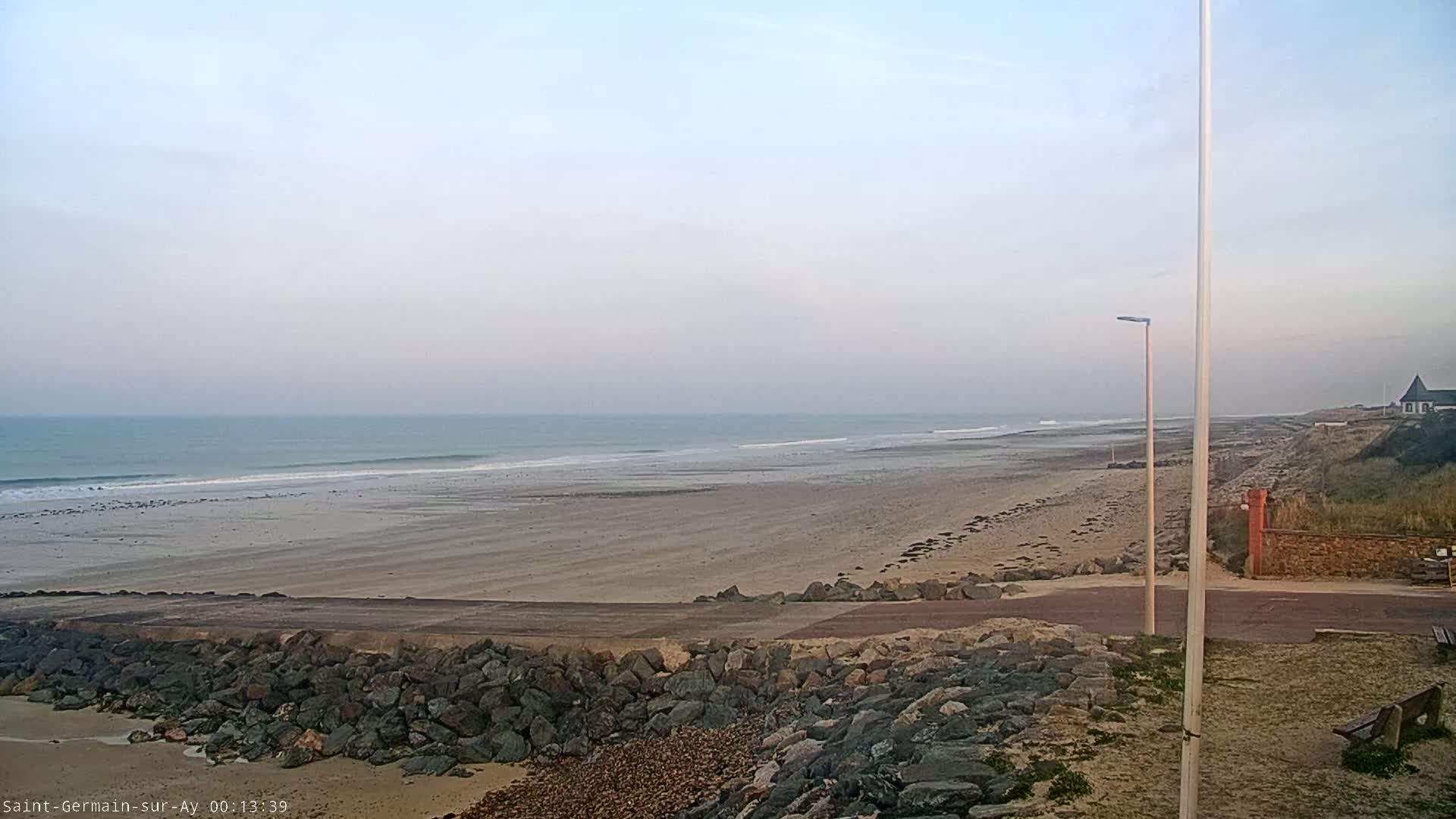 A wide sandy beach stretches to a calm ocean under a hazy sky, with a rocky embankment and a lamppost visible in the foreground.