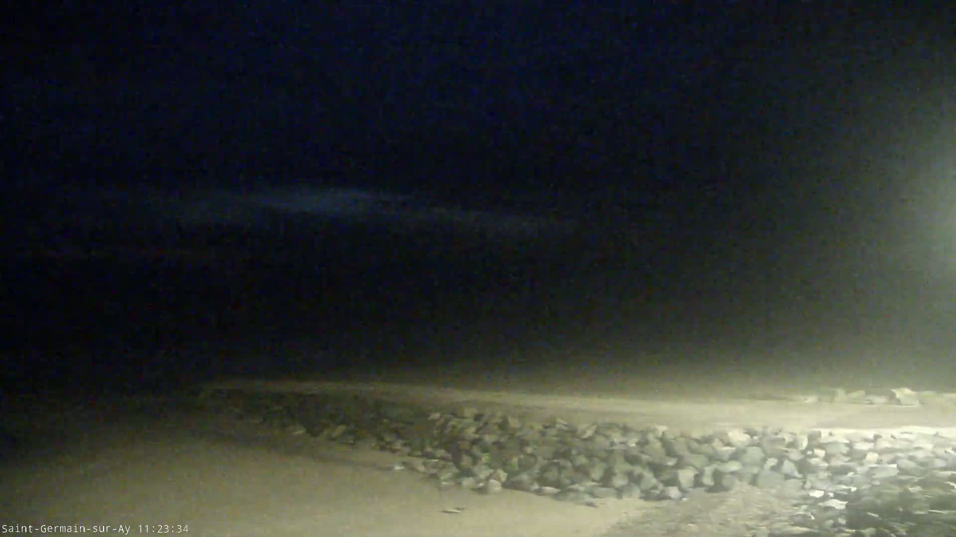 A dark, nighttime beach scene shows a low stone wall along the shore under a cloudy sky.