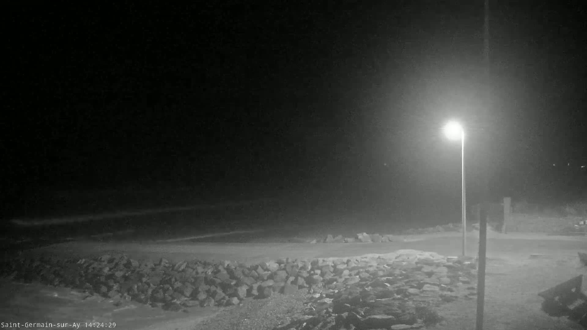 A nighttime view shows a rocky shoreline next to a dark body of water under a mostly clear sky, illuminated by a single lamppost.