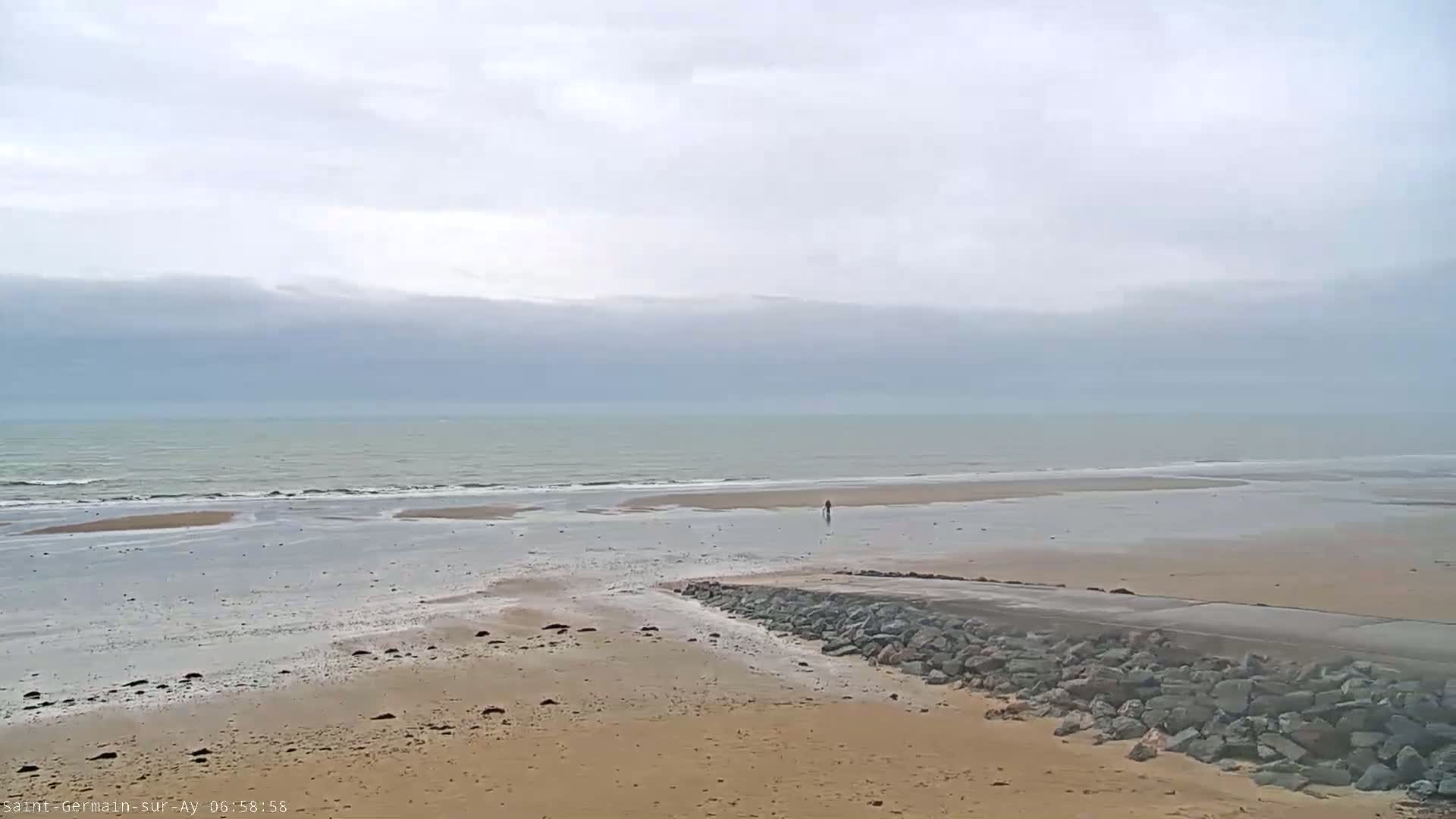 A wide sandy beach stretches to a calm, gray ocean under an overcast sky, with a single person visible in the distance.