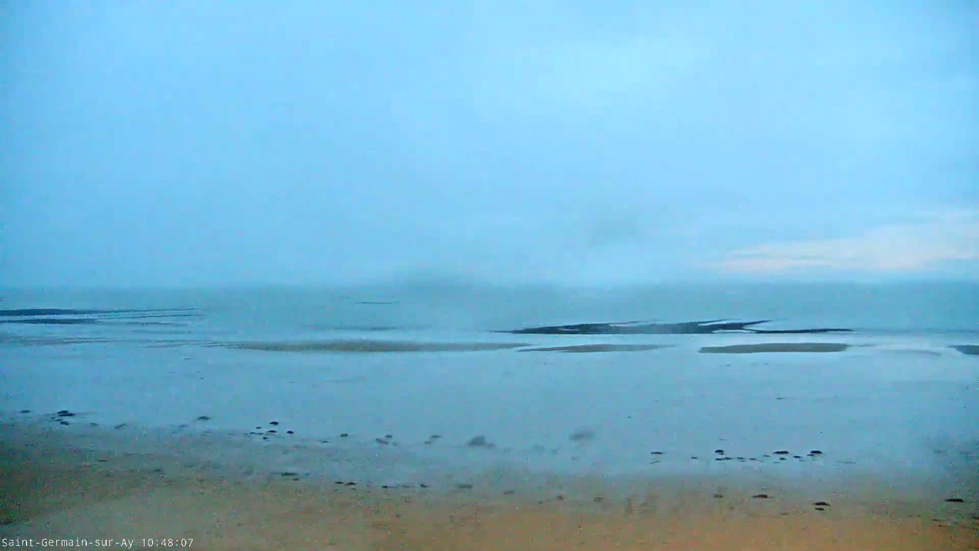 A hazy, overcast sky hangs over a calm, shallow body of water that meets a sandy beach.