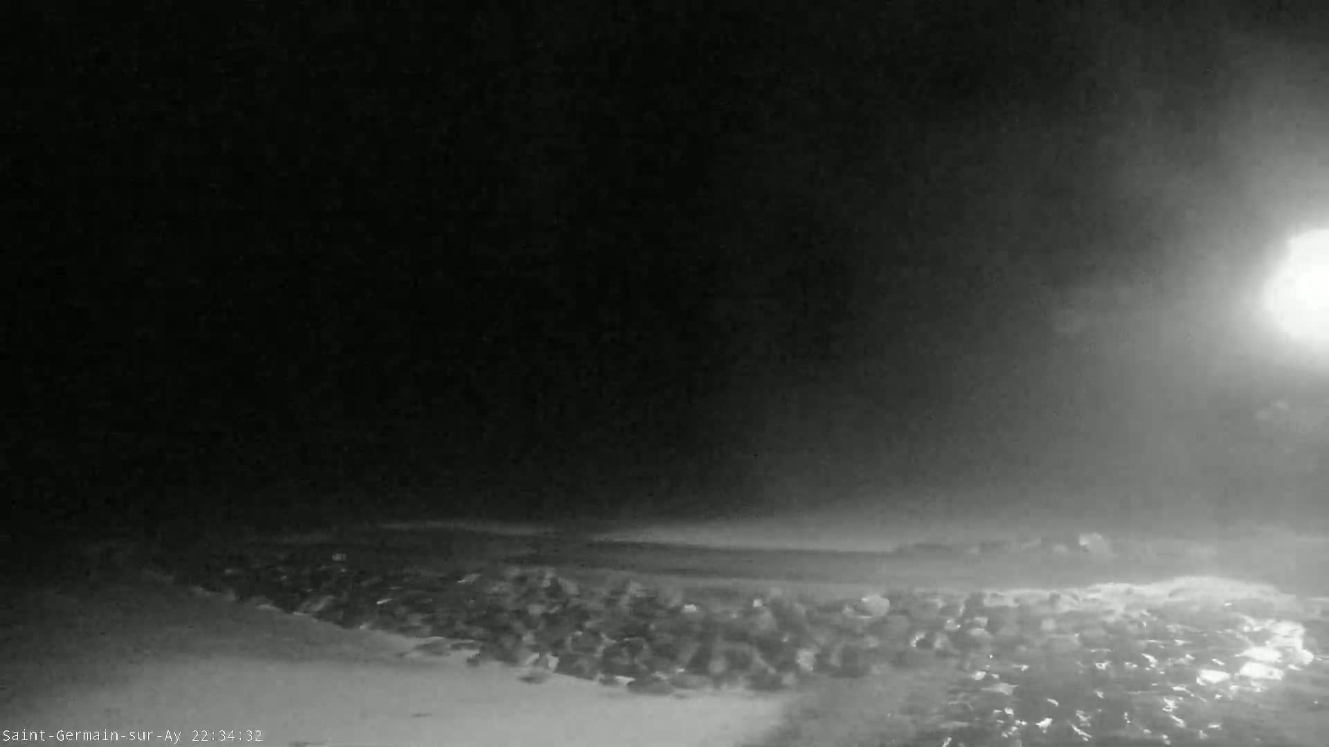 A nighttime, hazy scene shows a rocky shoreline under a dark sky with a bright light source in the upper right corner.