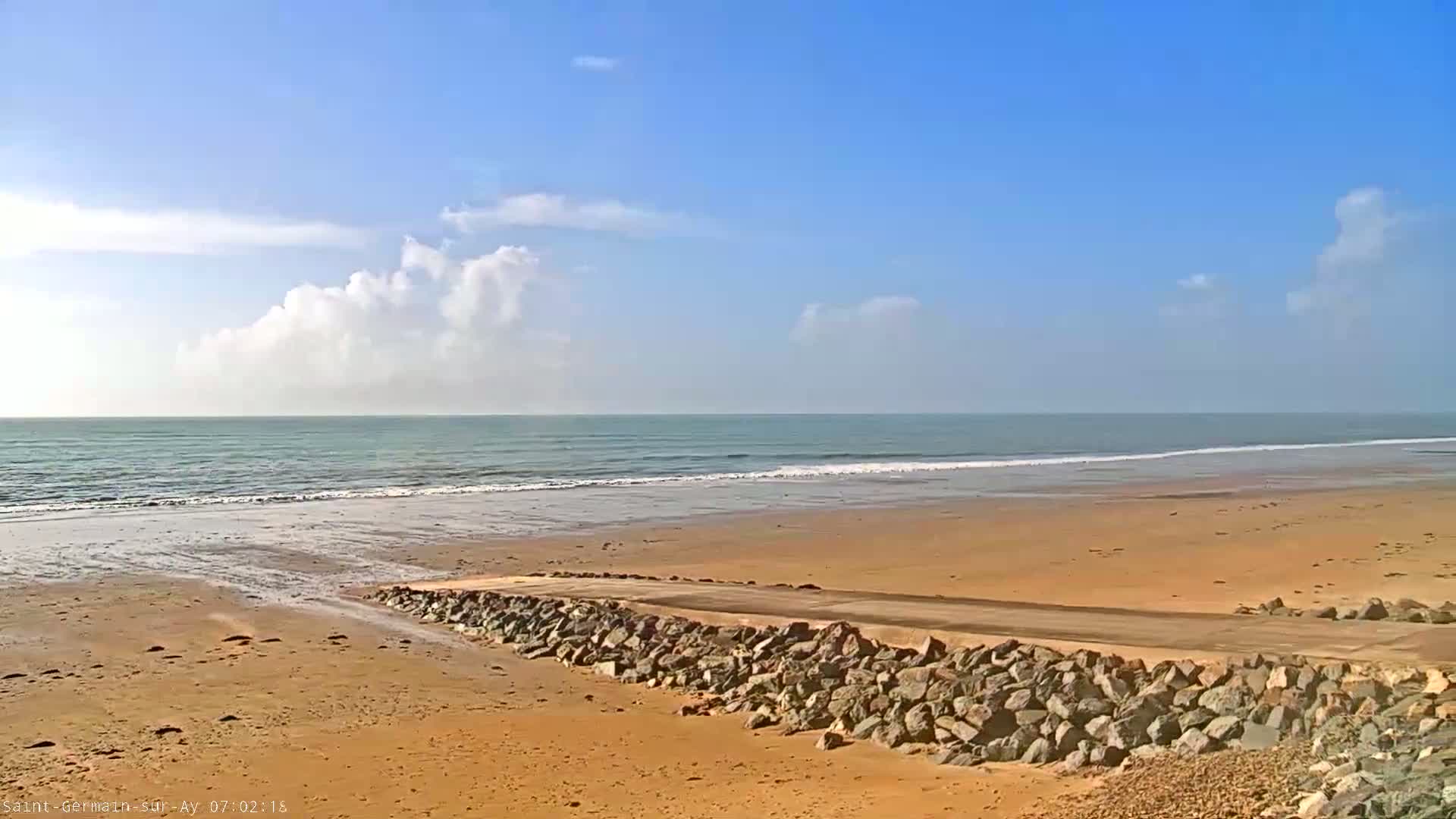 A sandy beach with a rock-lined road leading to the ocean under a mostly sunny sky with some cumulus clouds.