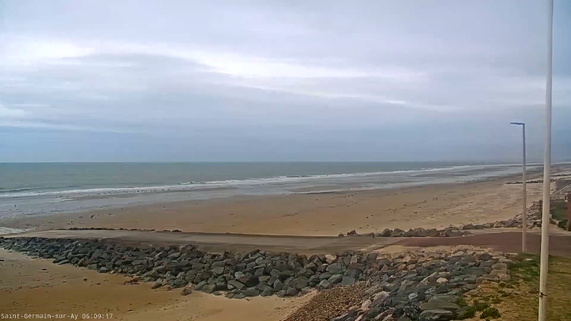 A wide sandy beach stretches to a calm ocean under a cloudy sky, with a rocky seawall and a lamppost visible in the foreground.