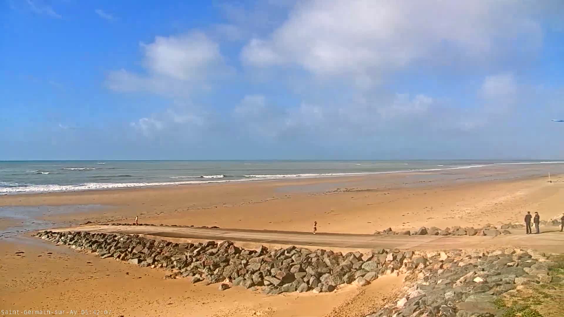 A sandy beach with a calm ocean under a partly cloudy blue sky features a paved road bordered by a rock wall, with several people standing near the road.