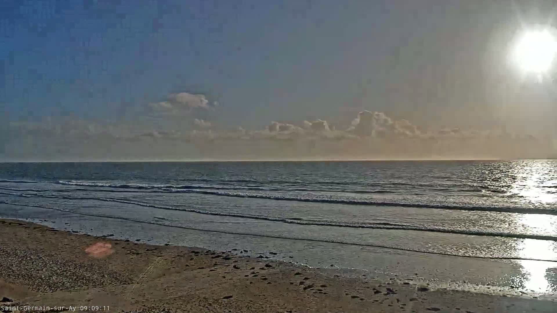 A sandy beach meets a calm ocean under a mostly clear sky with a bright sun.