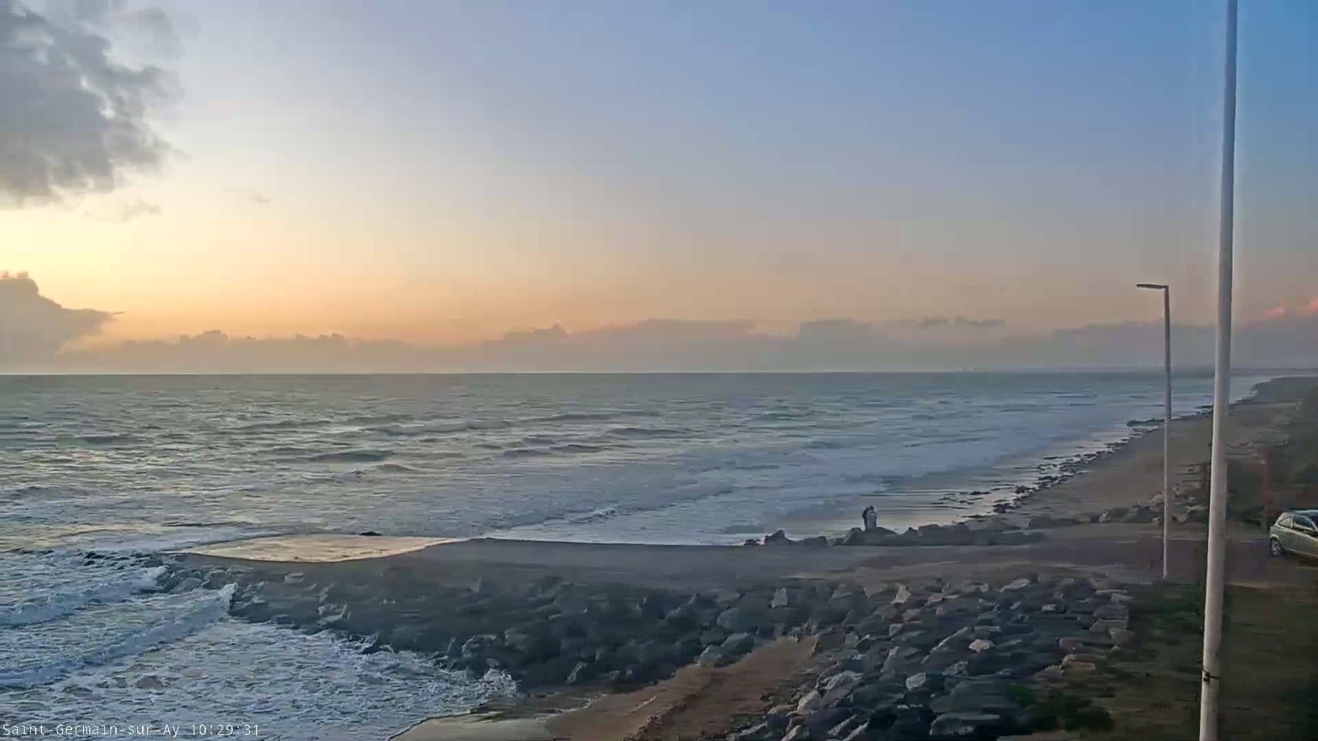 A calm ocean with small waves under a partly cloudy, sunrise sky meets a rocky shoreline with a few people standing near a vehicle.