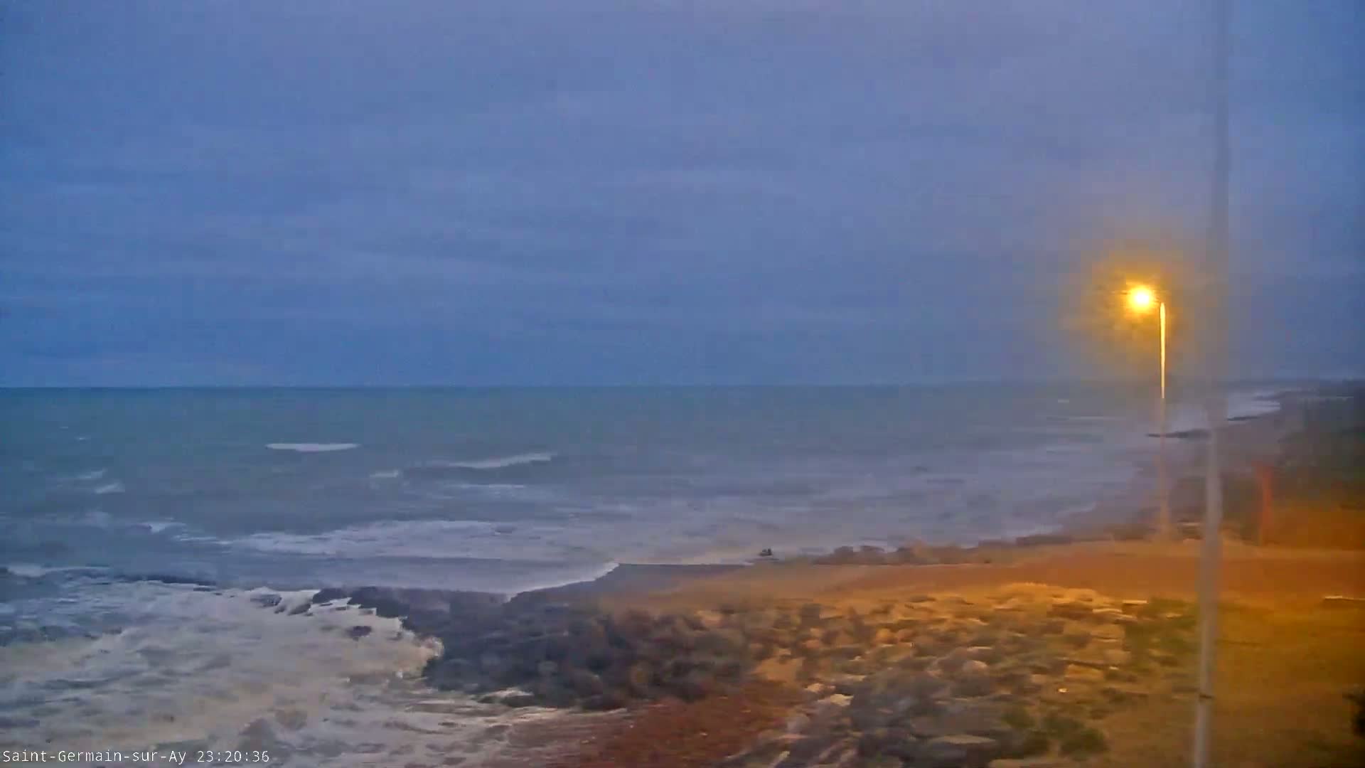 Under a dusky sky, moderately sized waves crash on a rocky shore next to a lit-up pathway.
