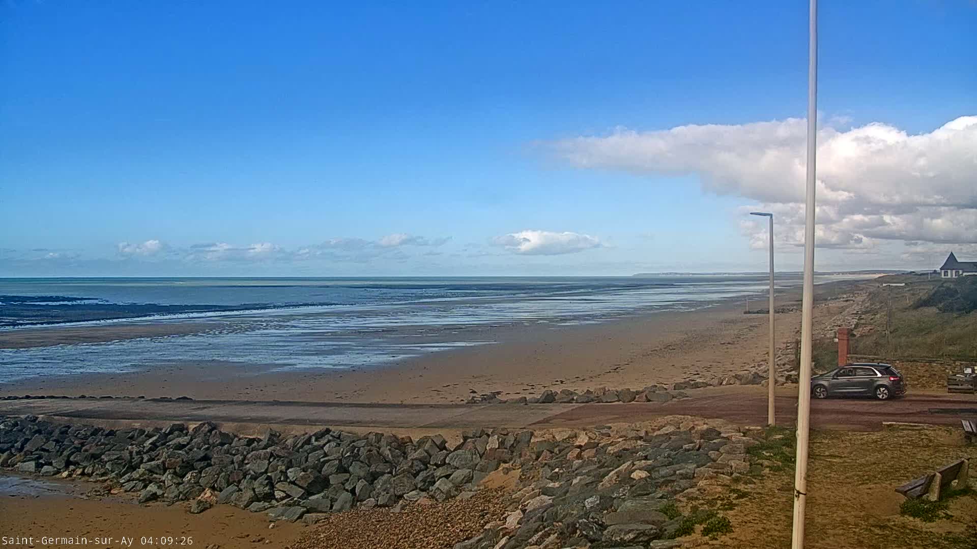 A sandy beach stretches to a calm ocean under a mostly clear blue sky with a few clouds, and a car is parked near a rocky embankment.