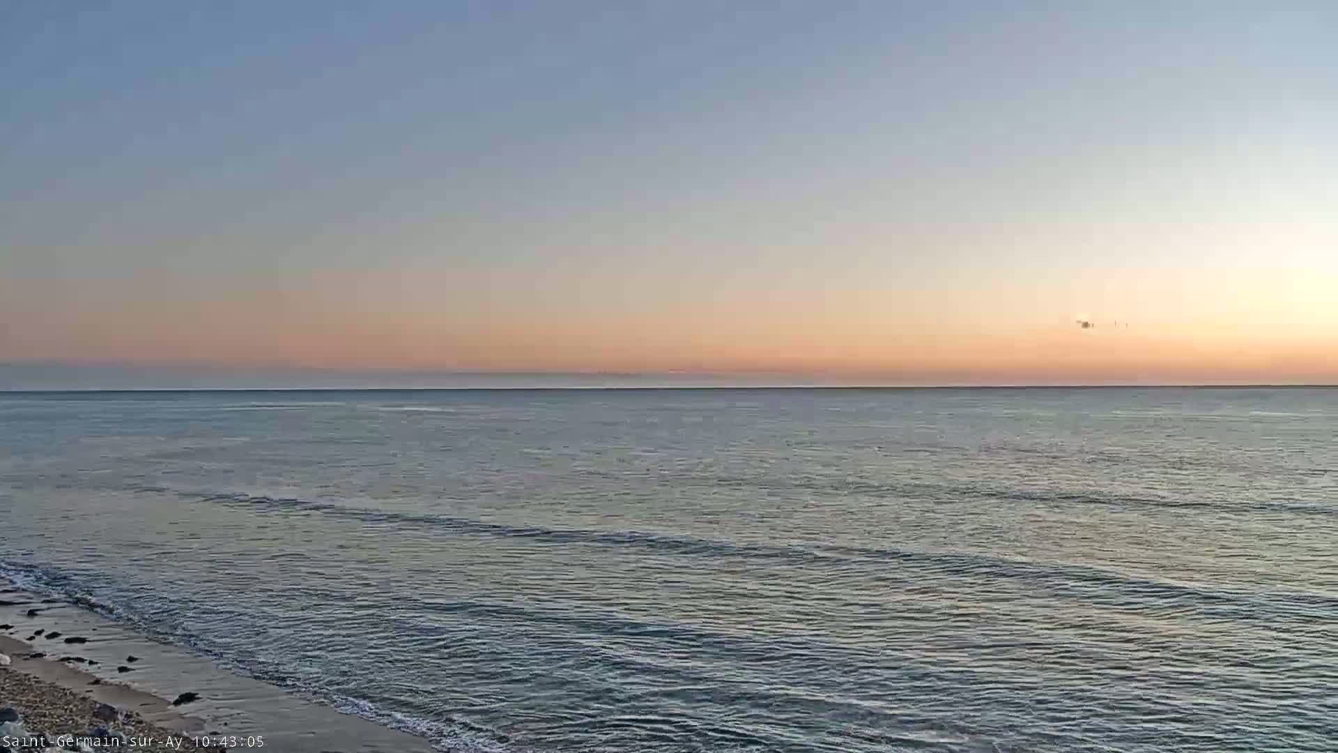 A calm ocean meets a pale pink and orange sunset sky over a pebbled shoreline.