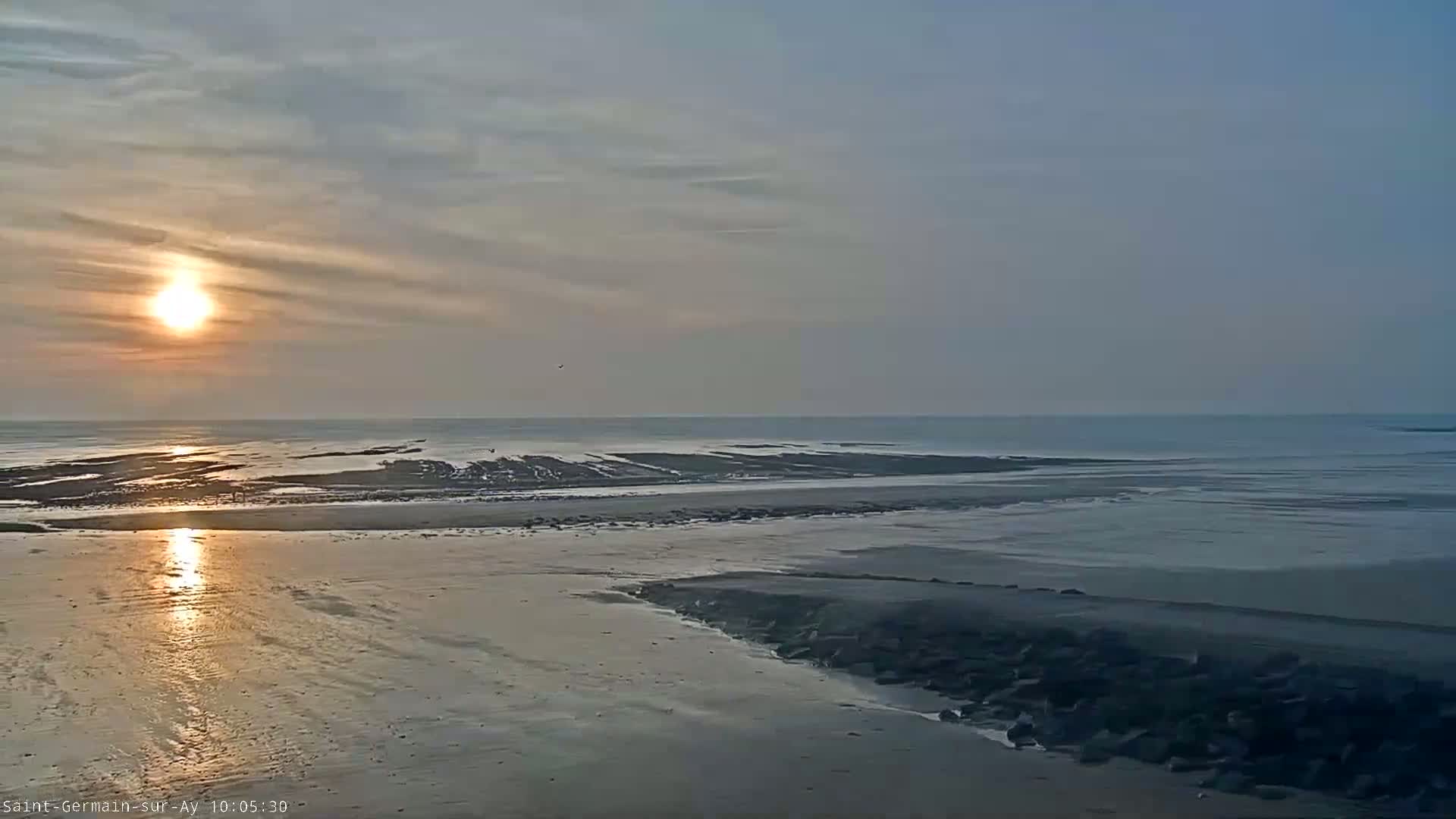 A low-tide beach at sunset shows a calm sea with a setting sun reflecting on the wet sand.