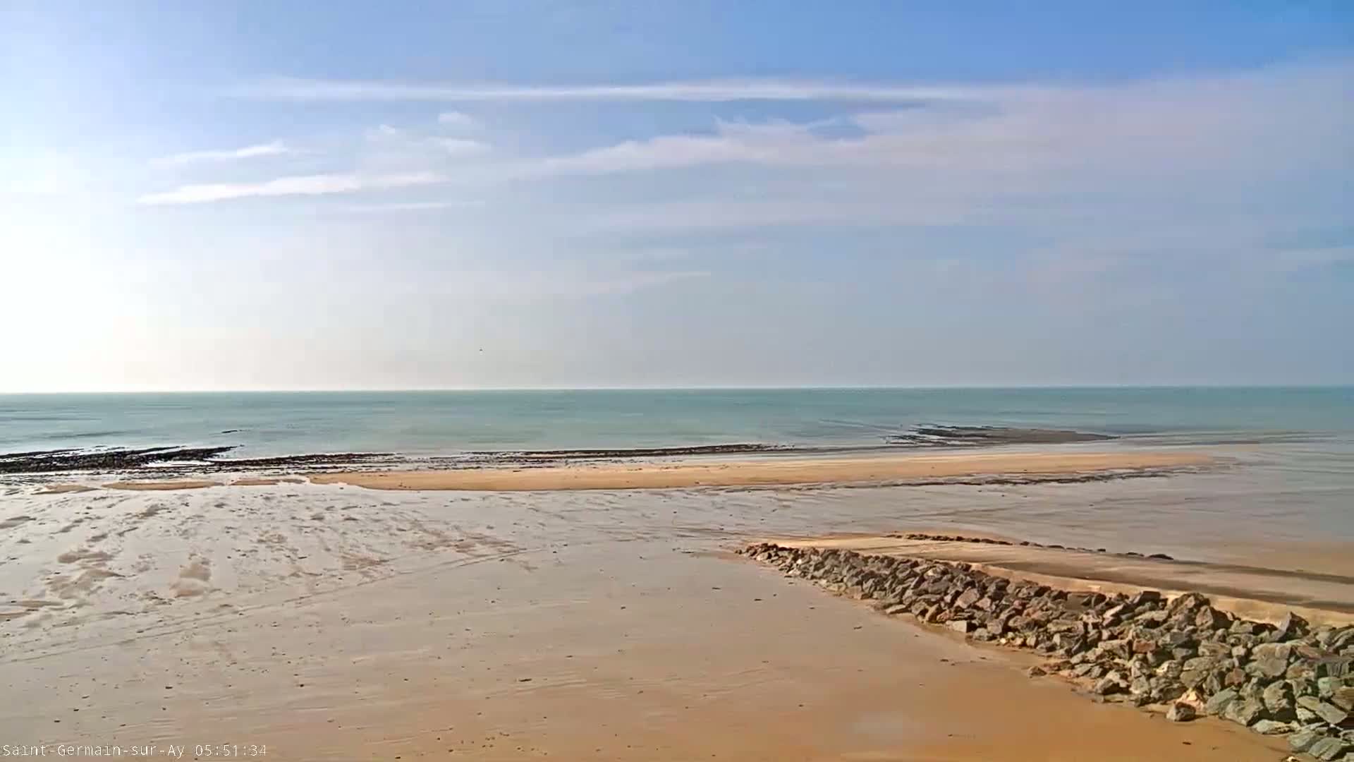 A mostly clear sky overlooks a calm, light-green ocean meeting a sandy beach with a low rock wall at the bottom right.