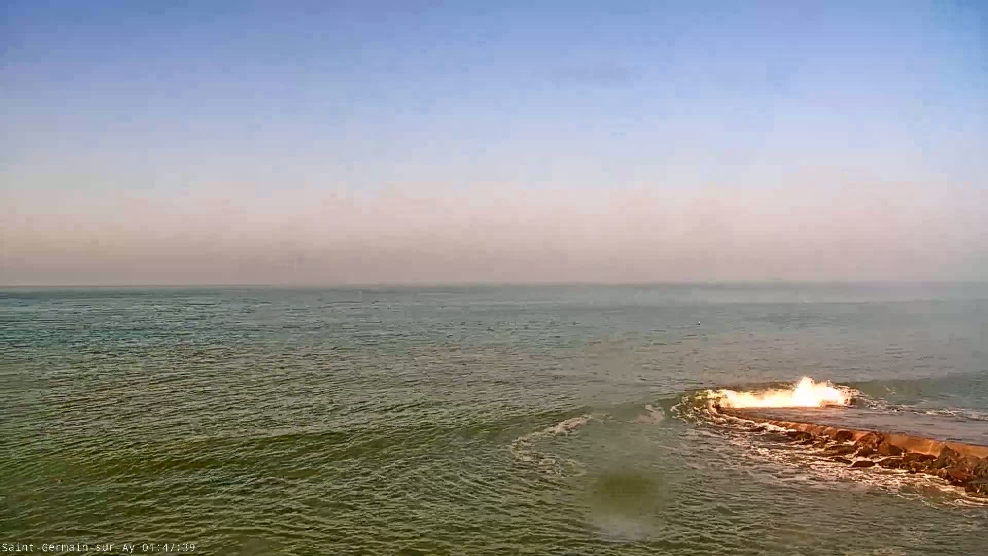Waves crash over a rocky outcropping in a hazy sea under a clear, pale sky.