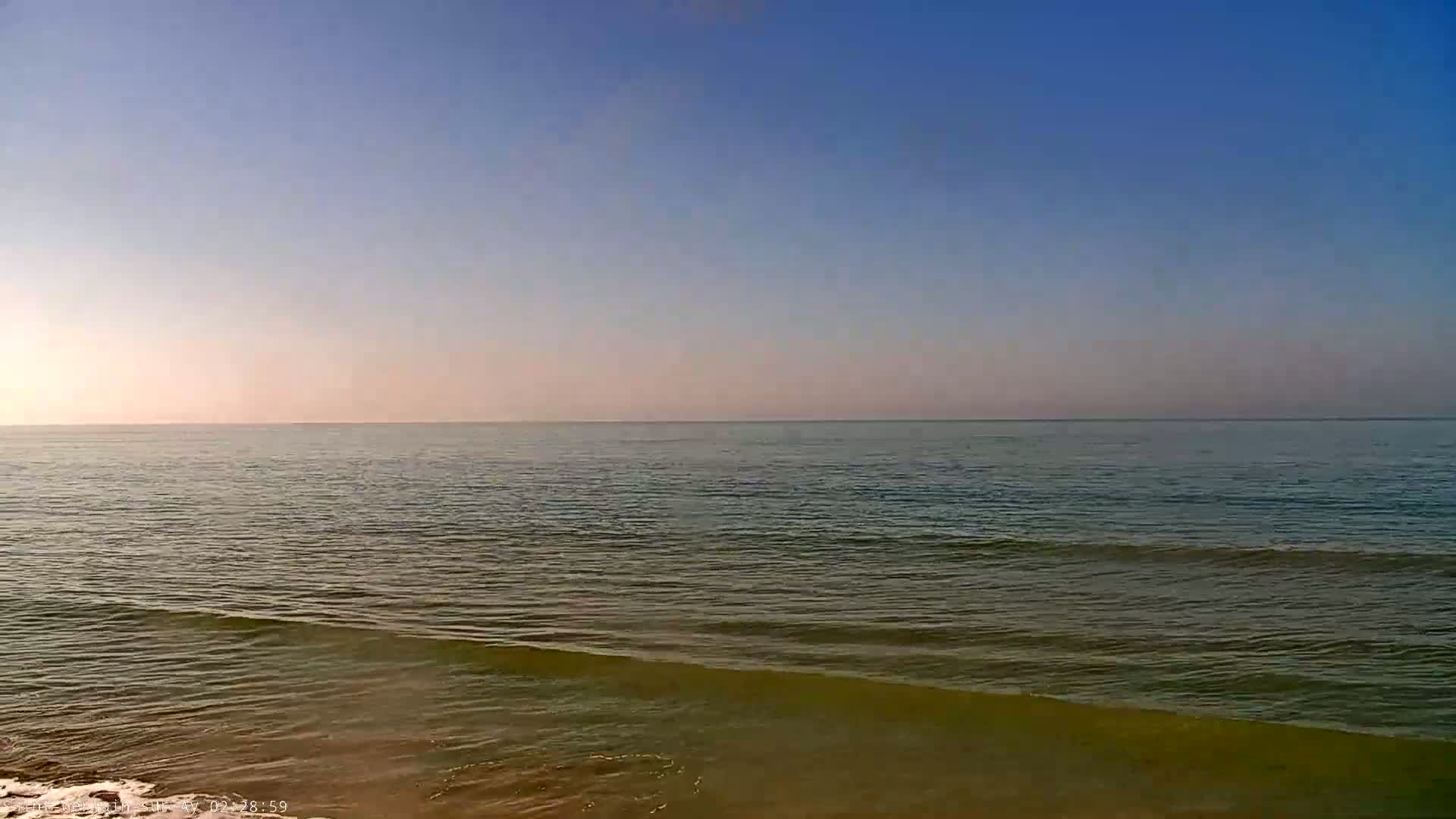 A calm sea with small waves laps a sandy shore under a clear, light-blue sky.