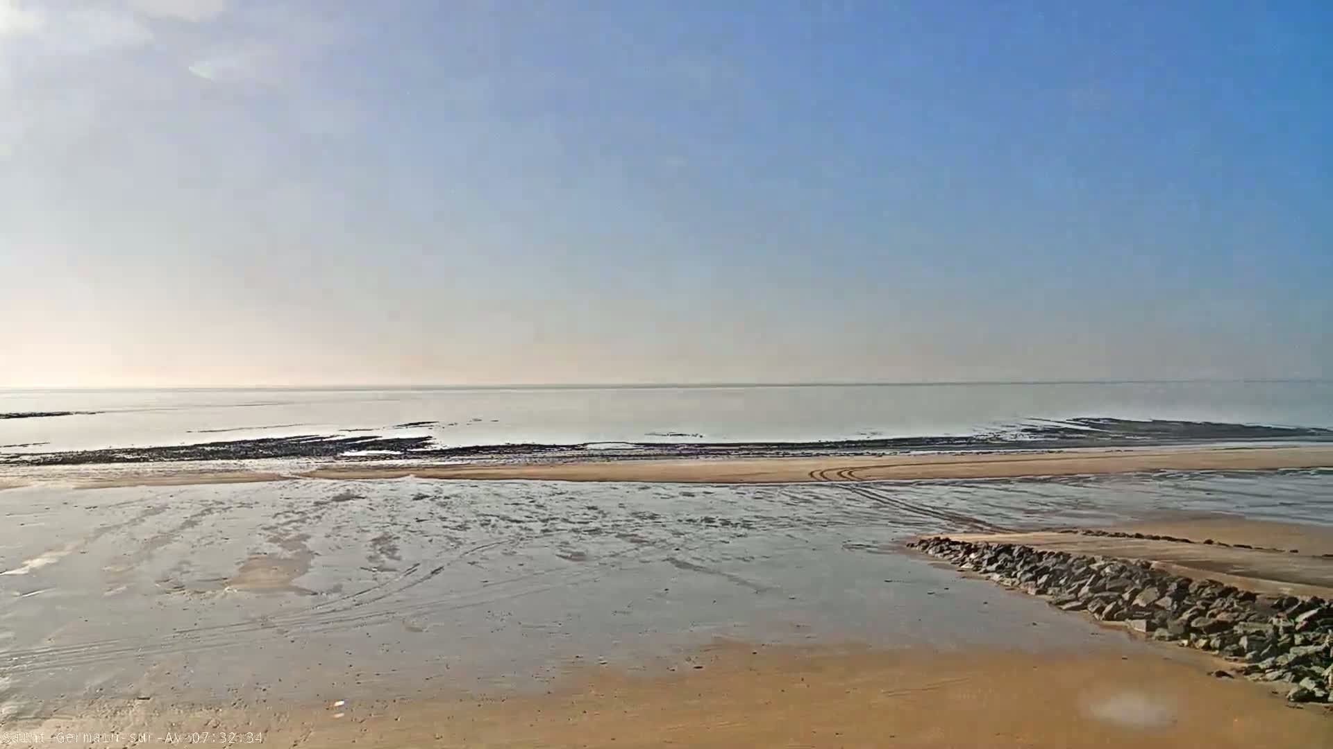 A sandy beach with tire tracks stretches toward a calm, light-blue ocean under a clear, bright sky.