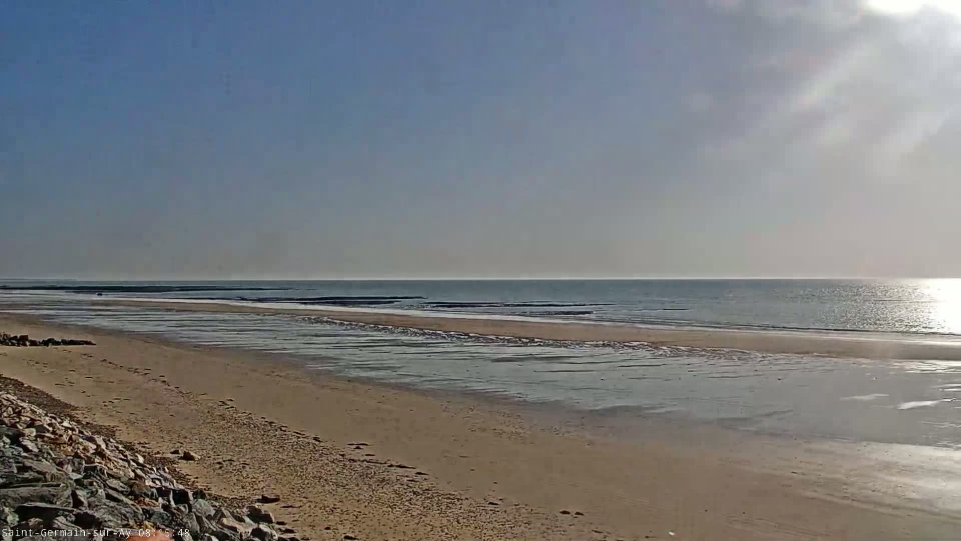 A sandy beach stretches to a calm, sunlit ocean under a mostly clear sky.