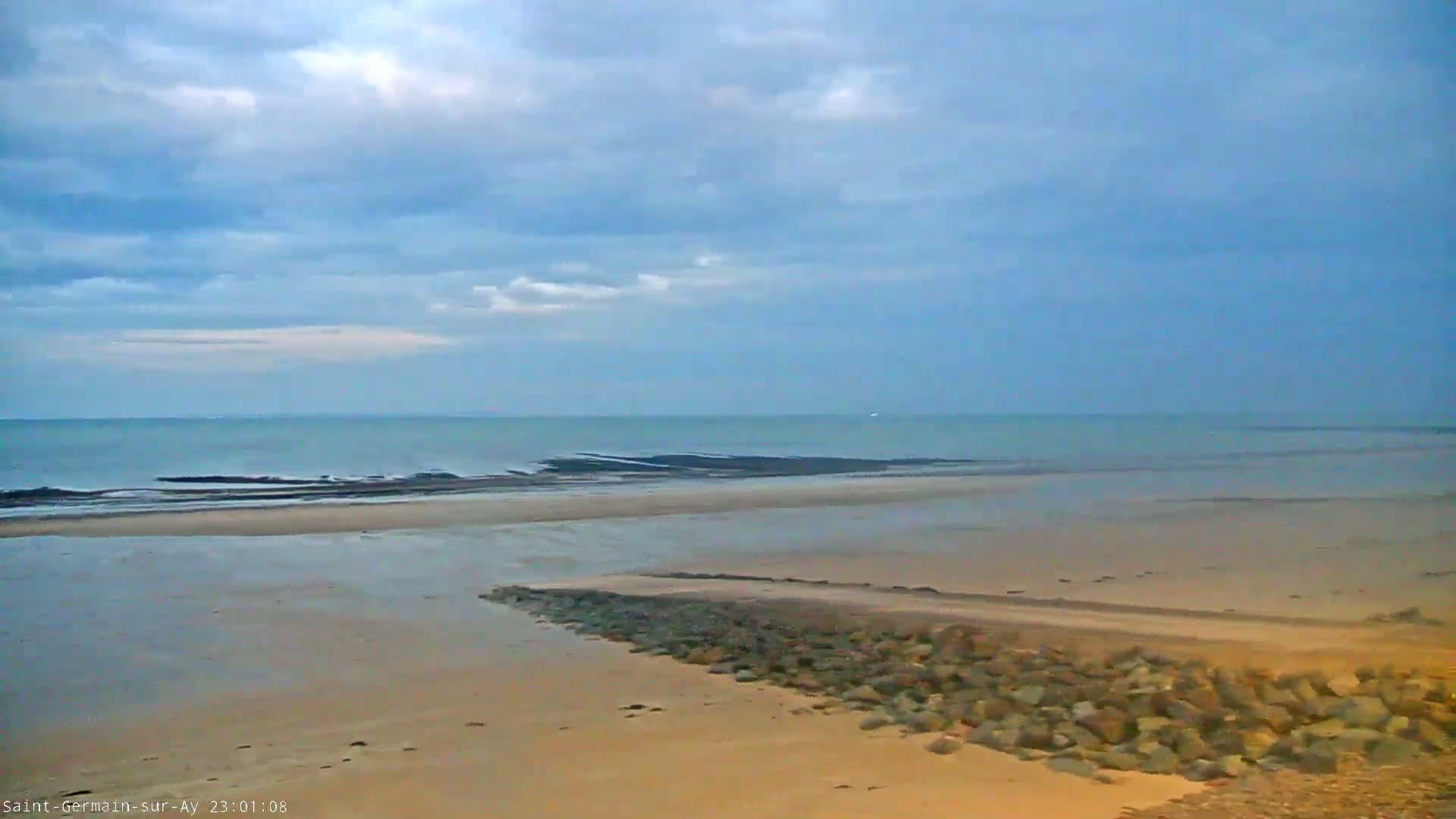 A sandy beach with a rocky breakwater stretches toward a calm sea under a mostly cloudy sky.