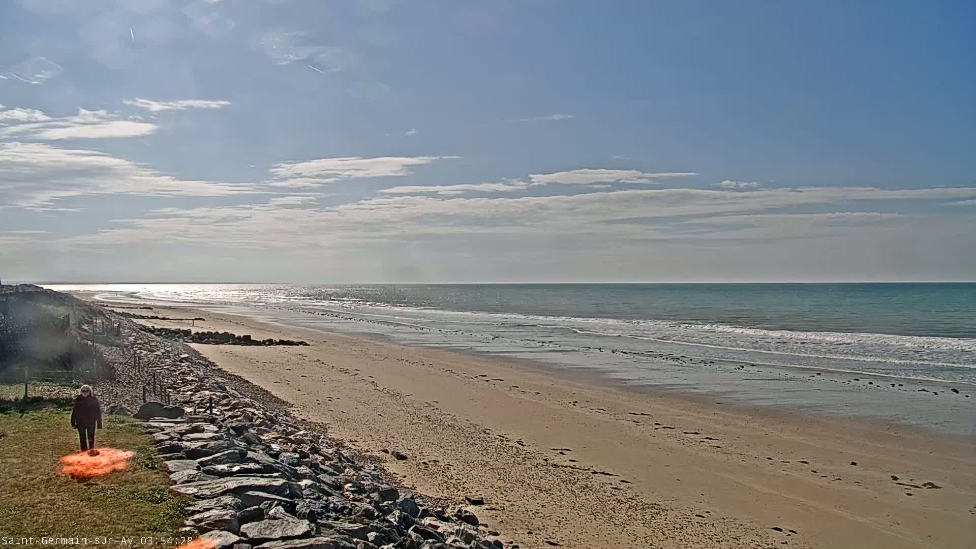 A person walks on grassy area next to a sandy beach with a calm ocean under a partly cloudy sky.