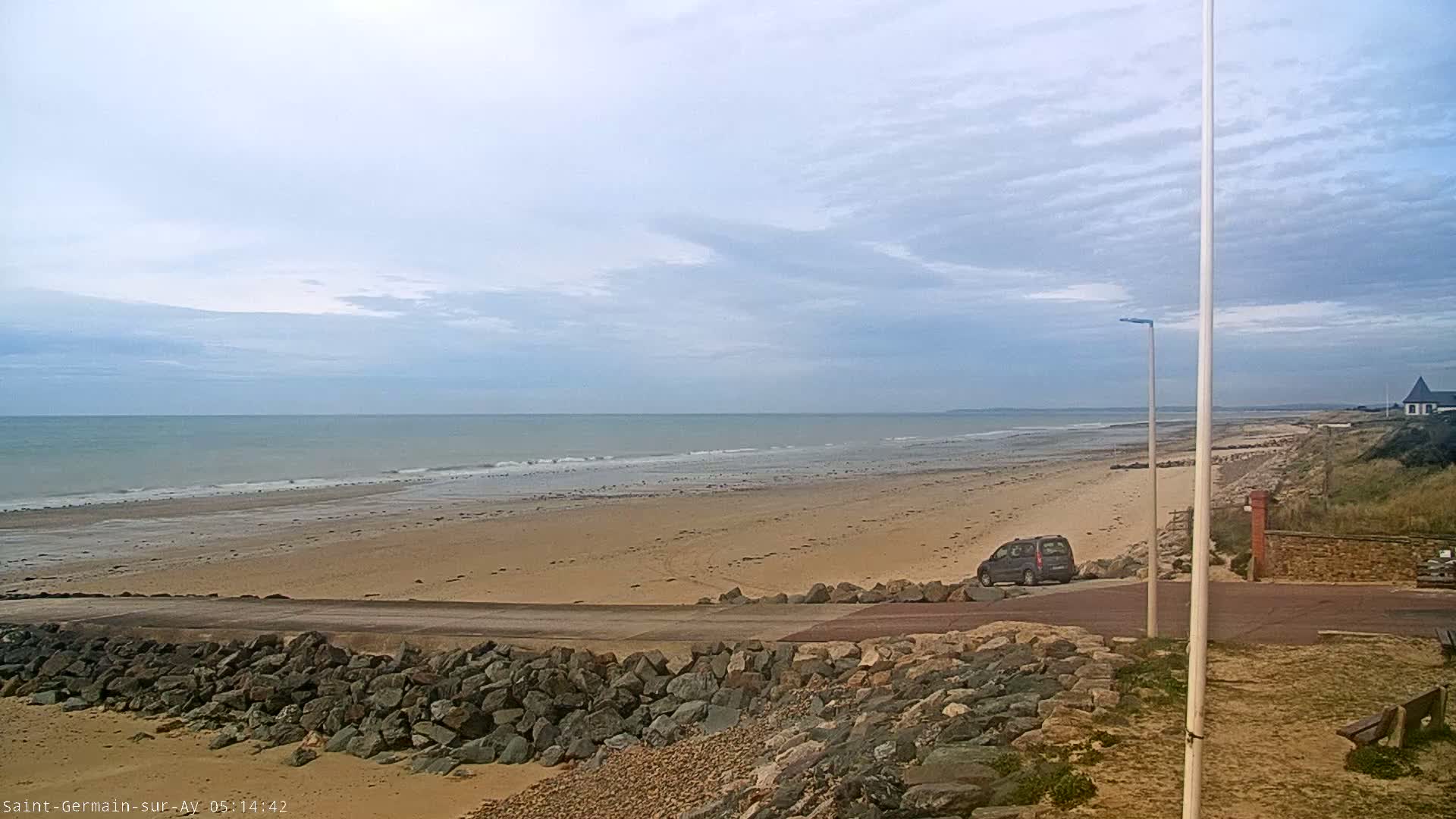 A sandy beach stretches to a calm ocean under a mostly cloudy sky, with a single vehicle parked near a rocky embankment.