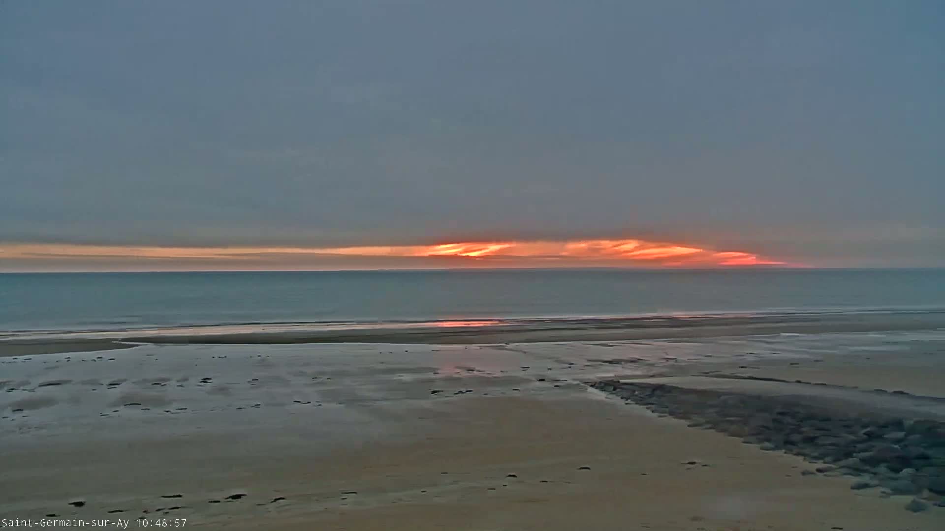 A calm ocean beach at sunset under a mostly cloudy sky displays a band of orange and pink hues on the horizon.
