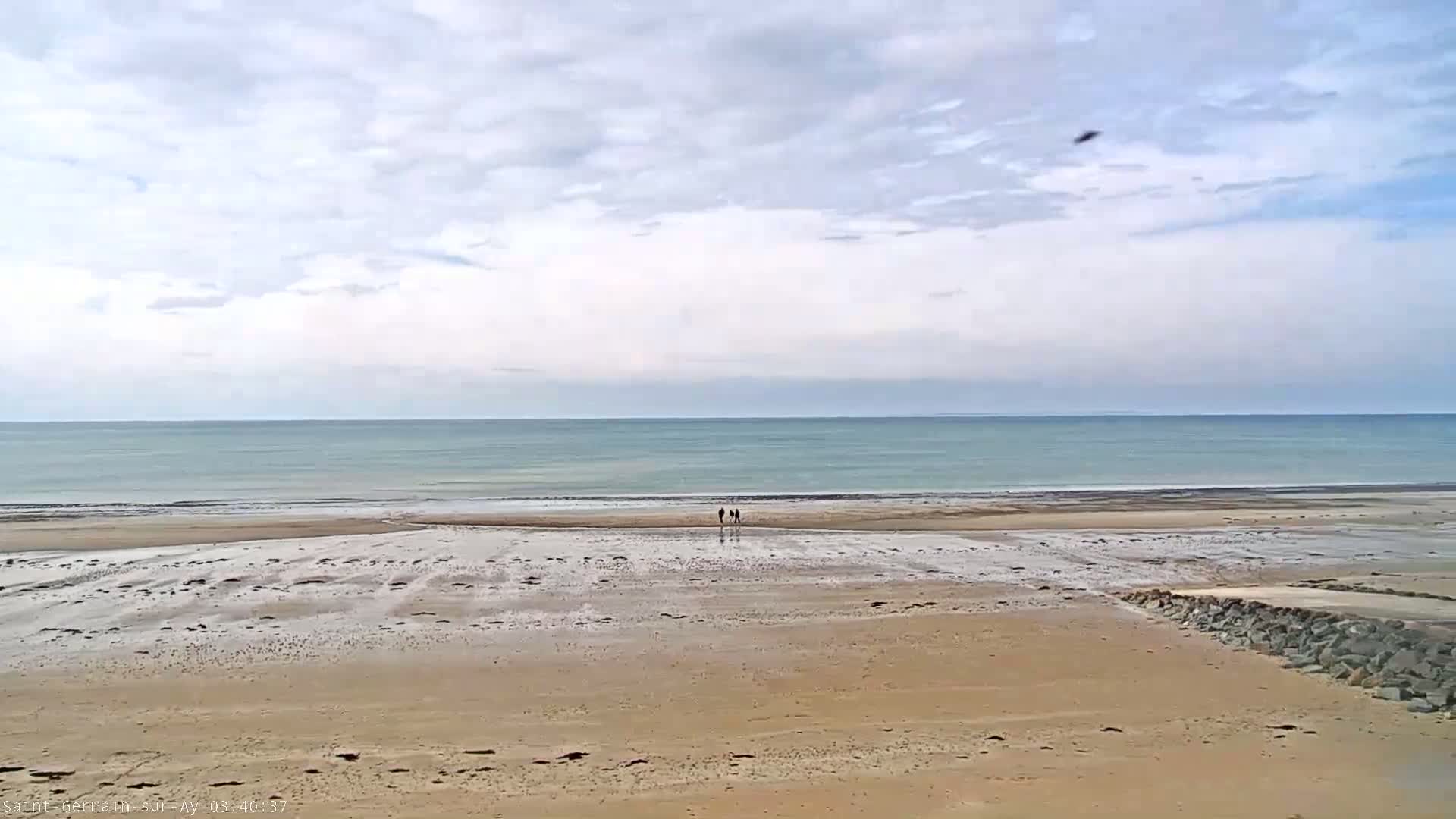 A wide sandy beach stretches toward a calm, light-blue ocean under a mostly cloudy sky, with a small group of people walking near the water's edge.