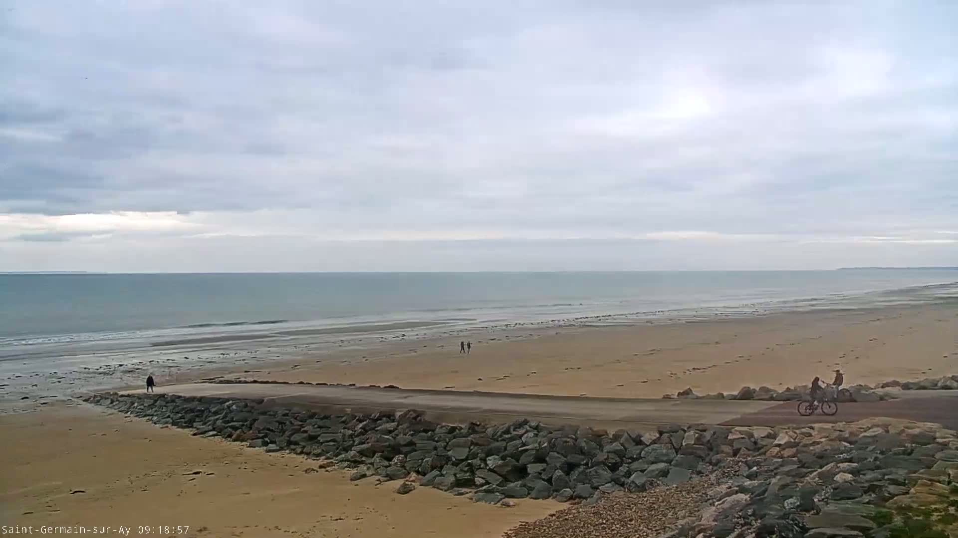 A wide sandy beach stretches to a calm ocean under an overcast sky, with a few people walking and biking on a paved path along the shoreline.