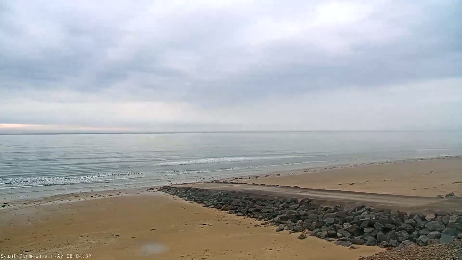 A sandy beach with a rock barrier and a calm, overcast ocean stretches to the horizon.