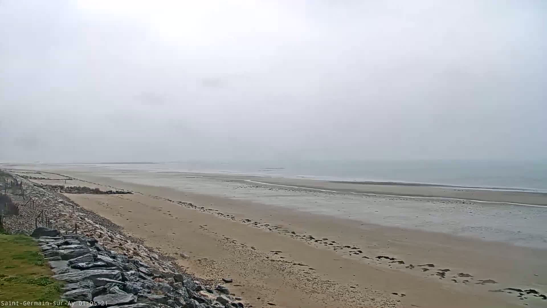 A wide, sandy beach stretches to a calm, gray sea under a foggy sky.