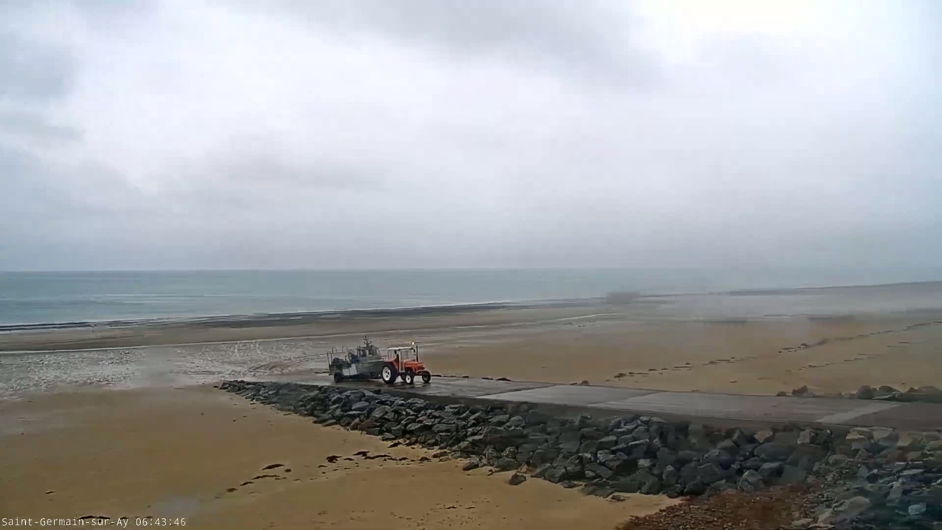 A tractor pulling a boat travels along a paved road that leads to a sandy beach and a calm, overcast ocean.