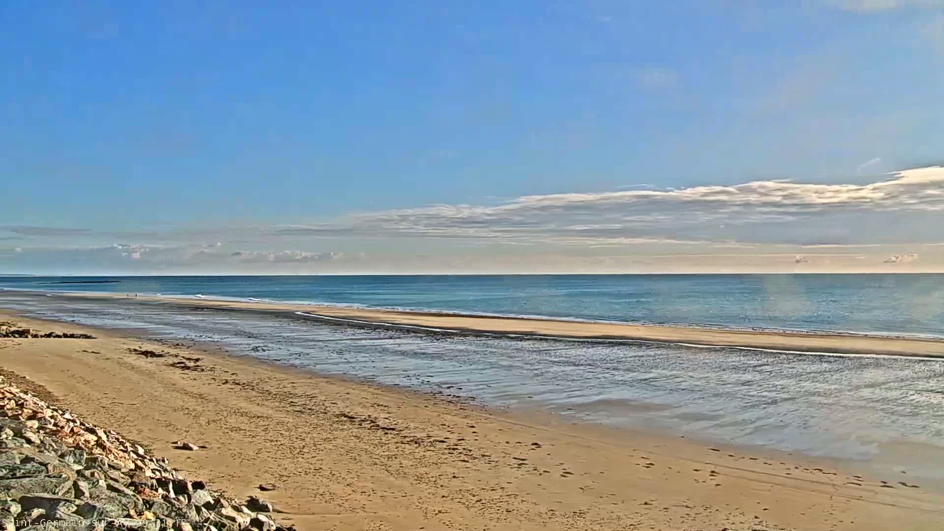 A sandy beach stretches to a calm, light-blue ocean under a mostly clear sky with scattered clouds.