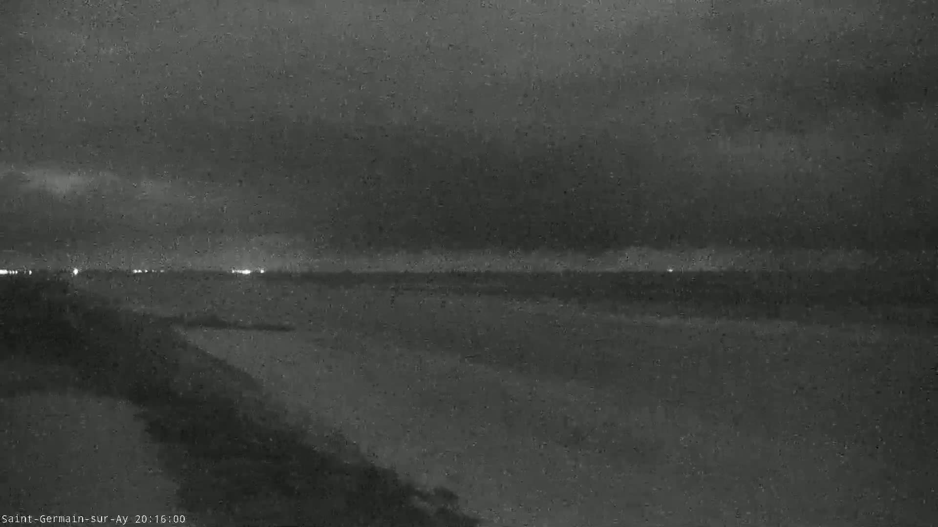 A nighttime view of a dark, possibly rainy, shoreline with faint lights visible in the distance.