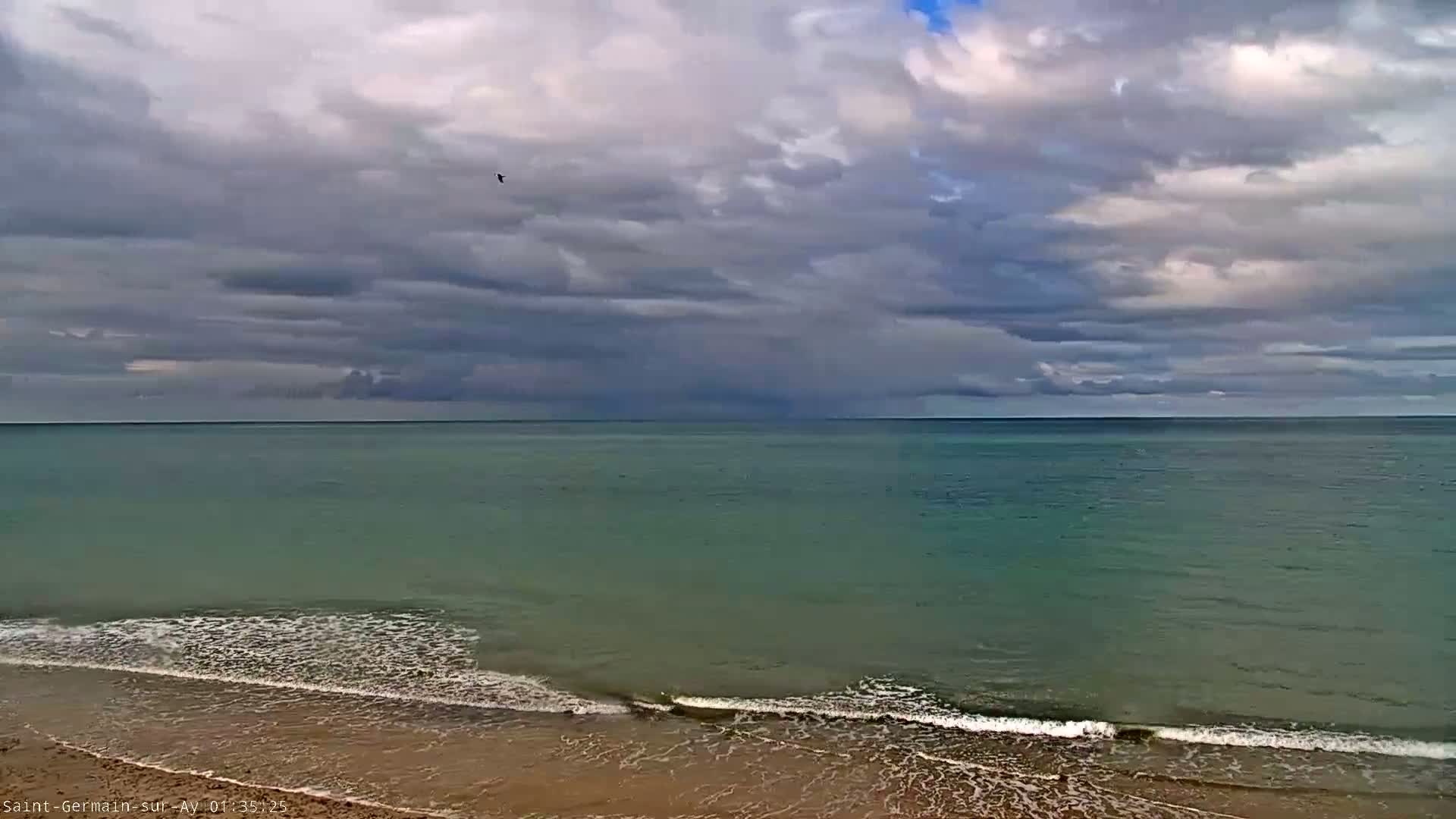 A mostly cloudy sky hangs over a calm, teal ocean meeting a sandy beach with small waves.