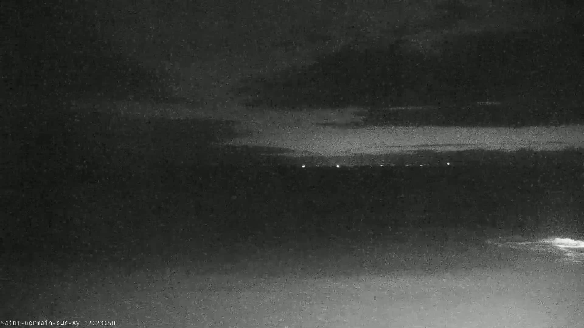 A nighttime, low-light view shows a dark landscape with a few distant lights and a dark body of water in the foreground.