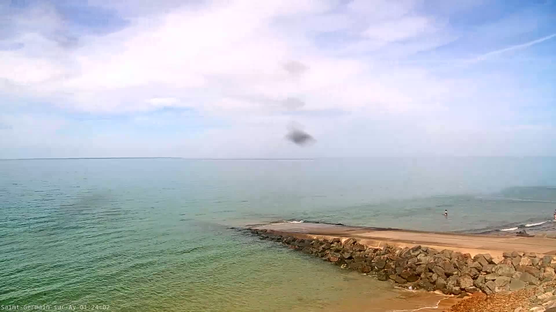 A rocky jetty extends into a calm, hazy ocean under a partly cloudy sky.