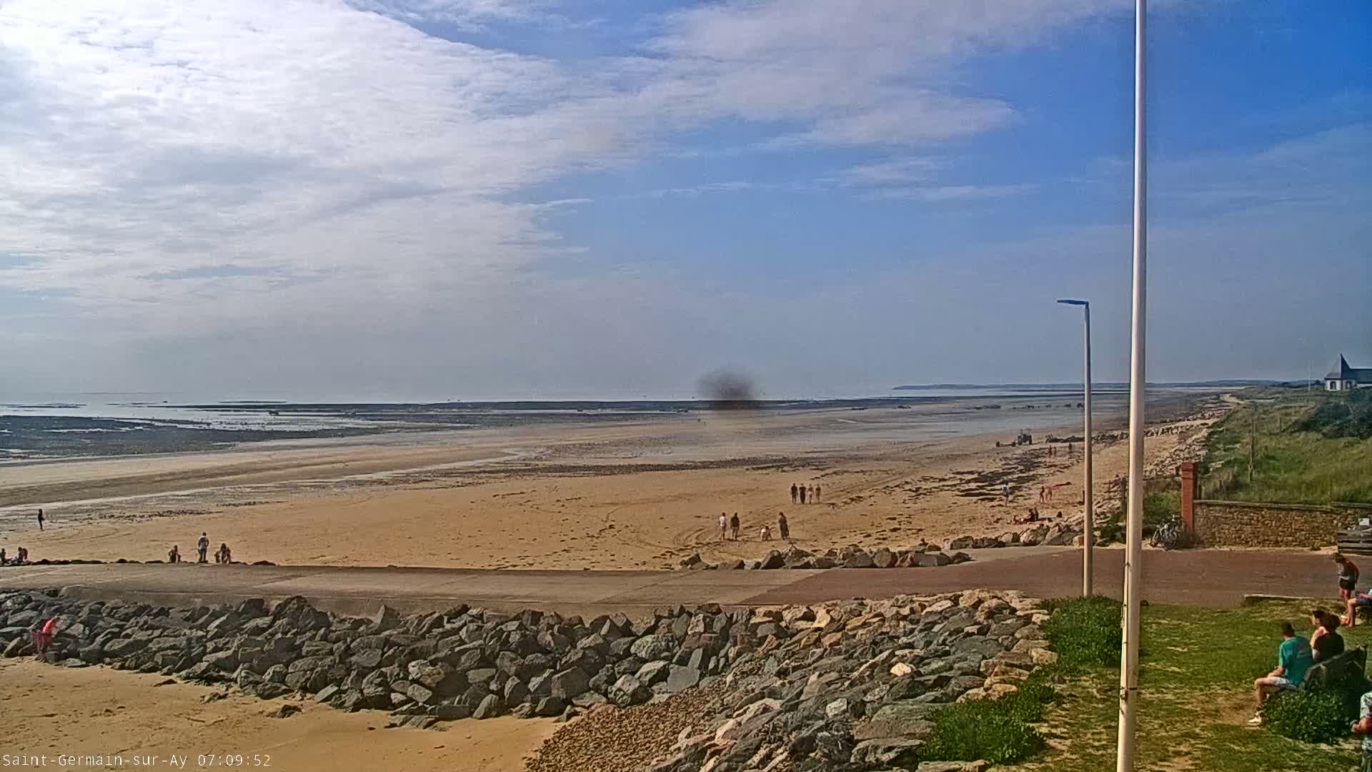 A sandy beach stretches to a calm ocean under a partly cloudy sky, with a few people scattered on the sand and others sitting near a rocky embankment.
