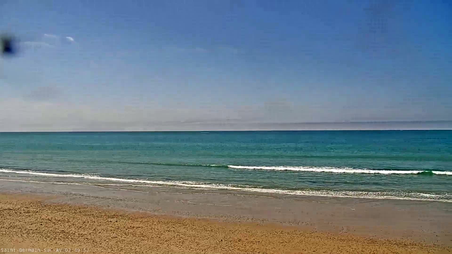 A sandy beach meets a calm, teal ocean under a clear, mostly sunny sky.