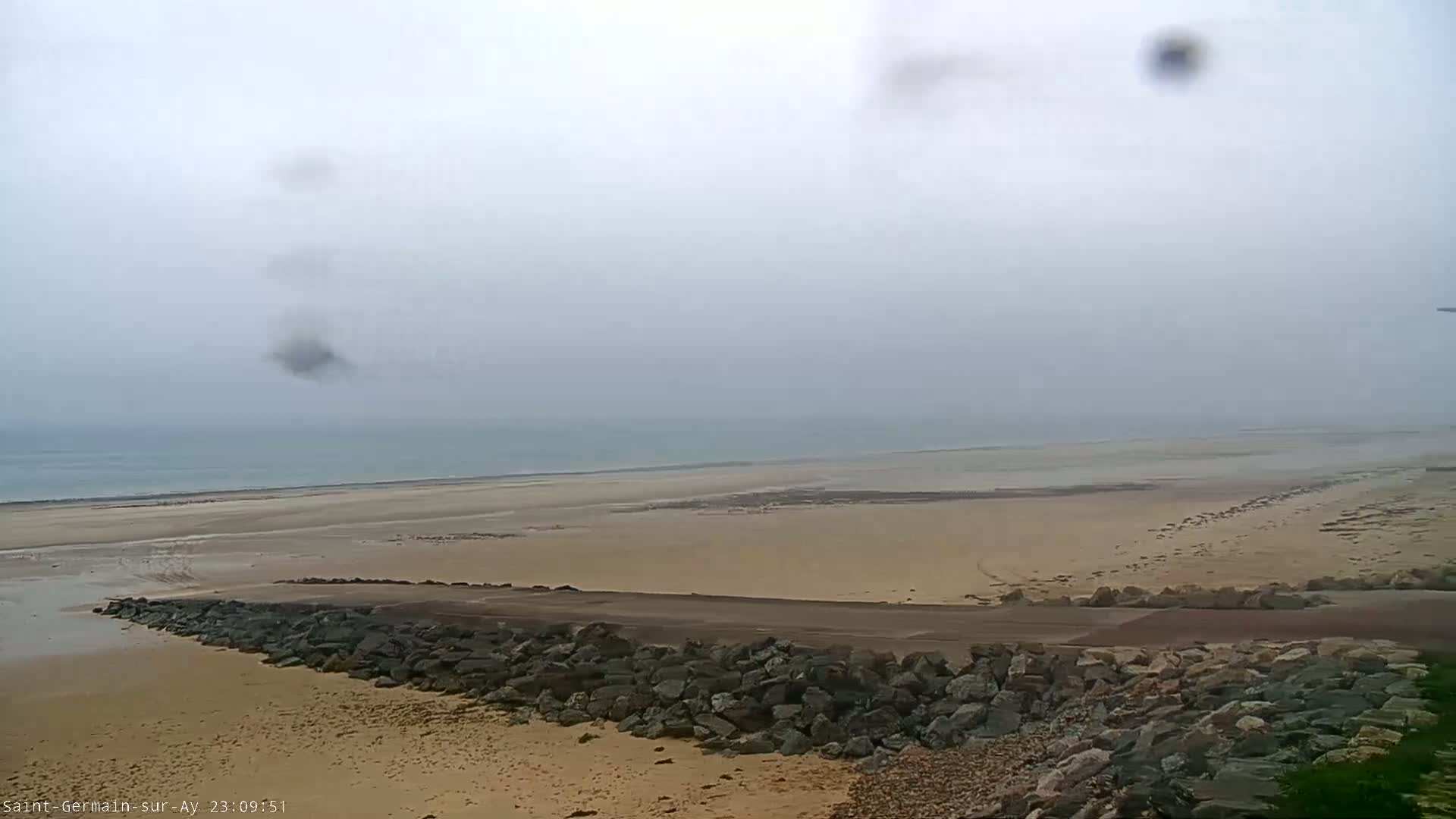 A foggy day reveals a wide expanse of sandy beach meeting a calm sea, partially obscured by a rocky embankment.