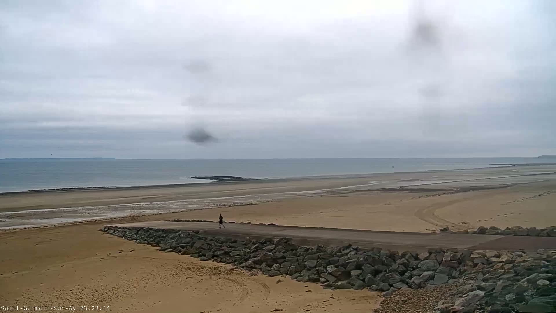 A person jogs along a paved path that separates a sandy beach from a calm, gray-sky ocean.