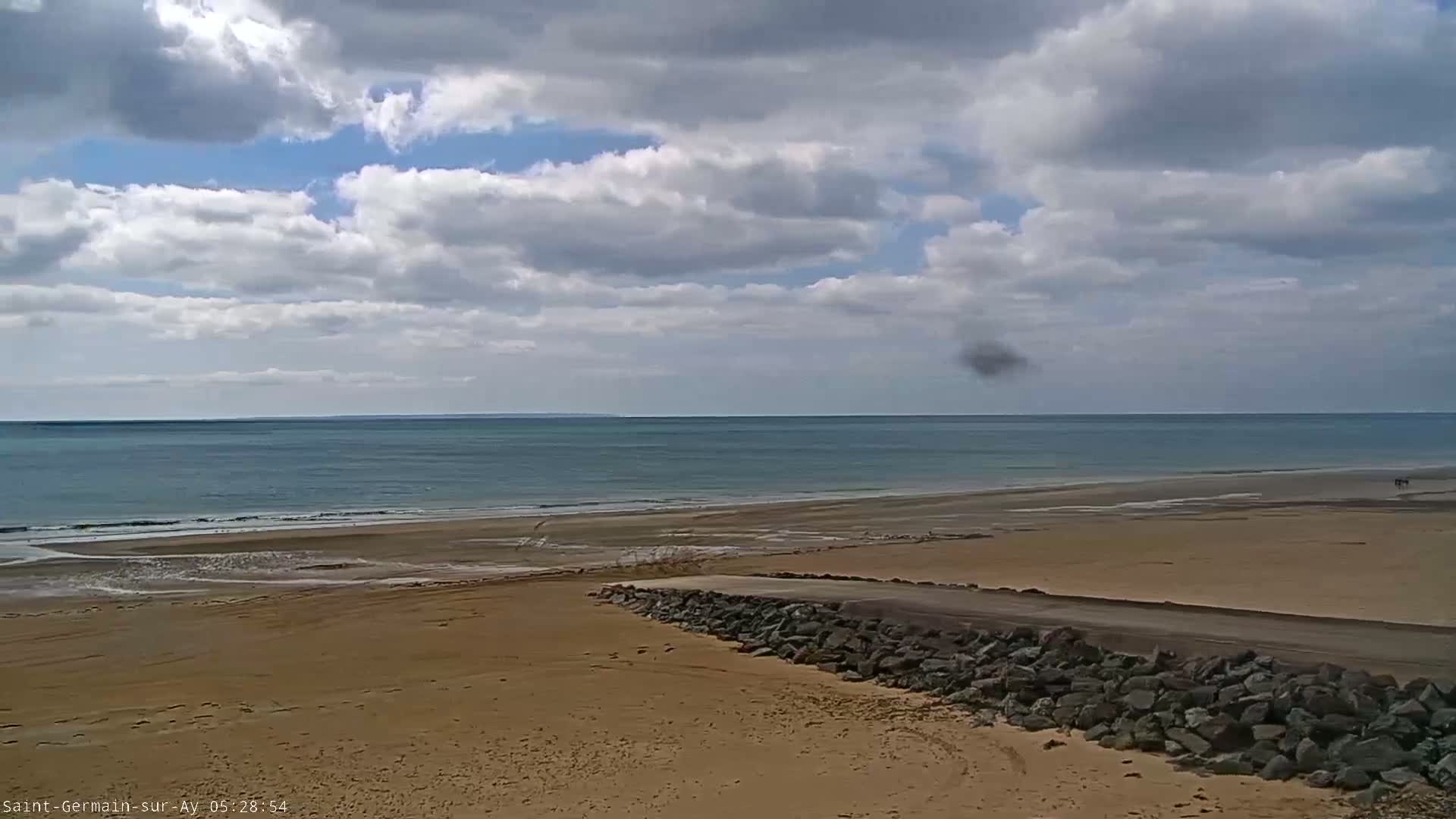 A partly cloudy day shows a wide sandy beach meeting a calm ocean with a small rock wall separating the beach from a roadway.