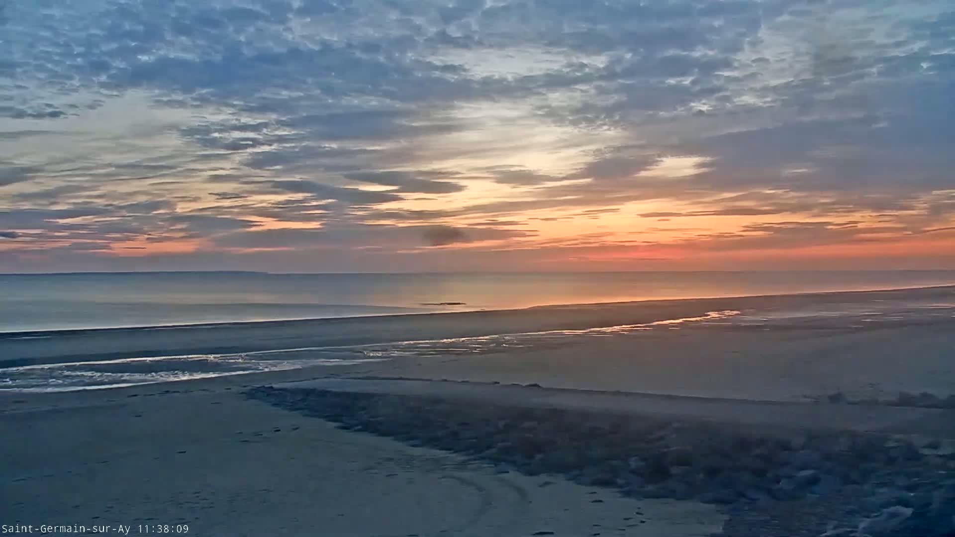 A tranquil beach scene at sunset displays a calm ocean under a mostly cloudy, pinkish-orange sky.