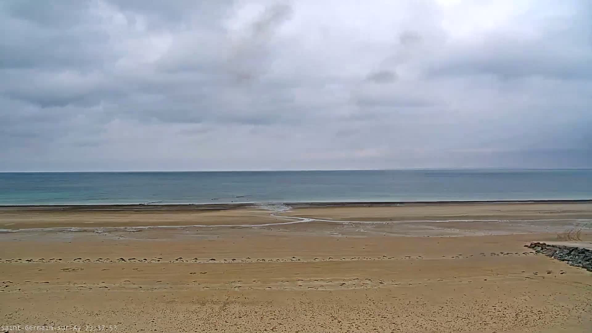 A wide expanse of sandy beach stretches to a calm, pale-blue ocean under a cloudy sky.