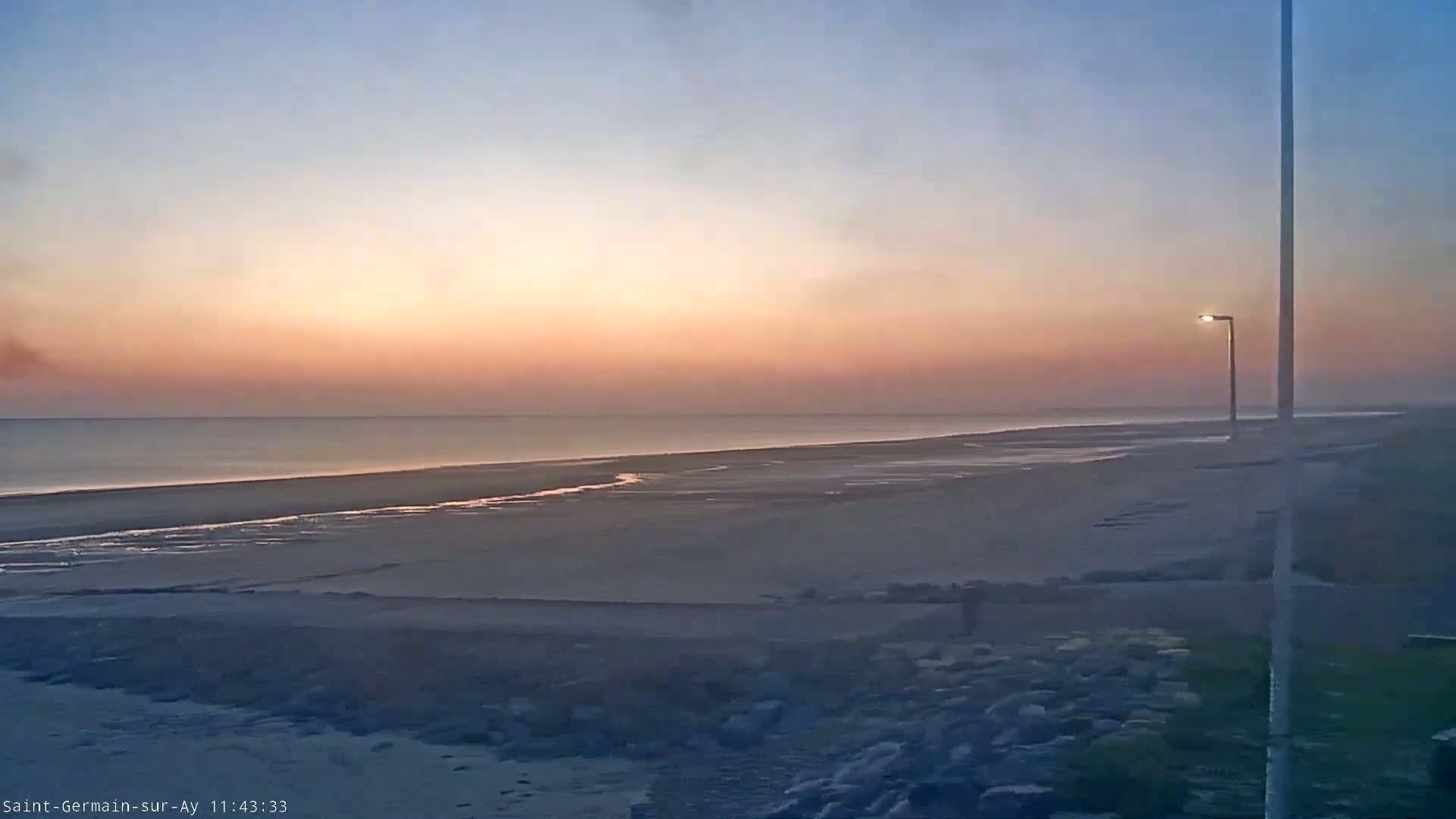 A calm ocean meets a wide, sandy beach at sunset under a pale pink and orange sky, with a single lamppost standing on the shoreline.