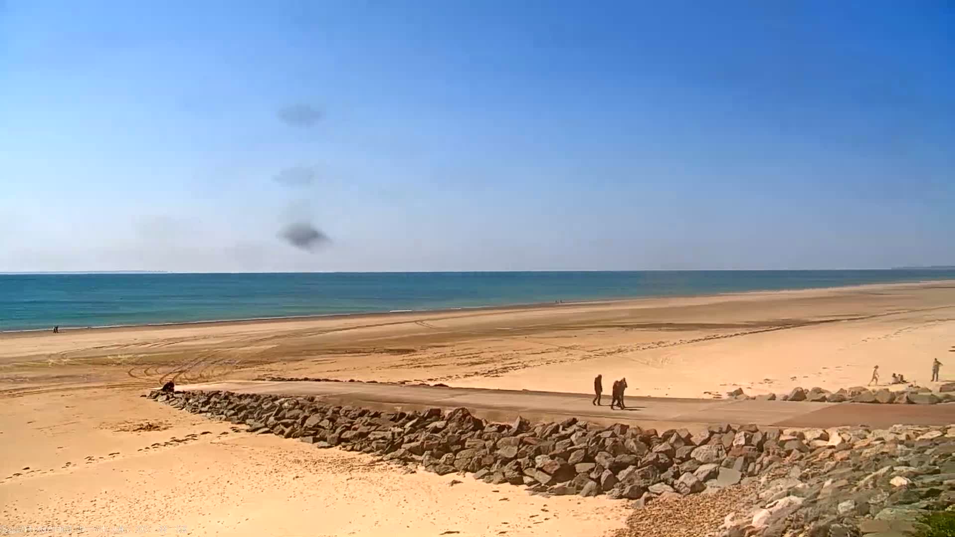 A wide expanse of sandy beach stretches to a calm blue ocean under a clear blue sky, with a few people walking along a paved path that cuts across the beach.