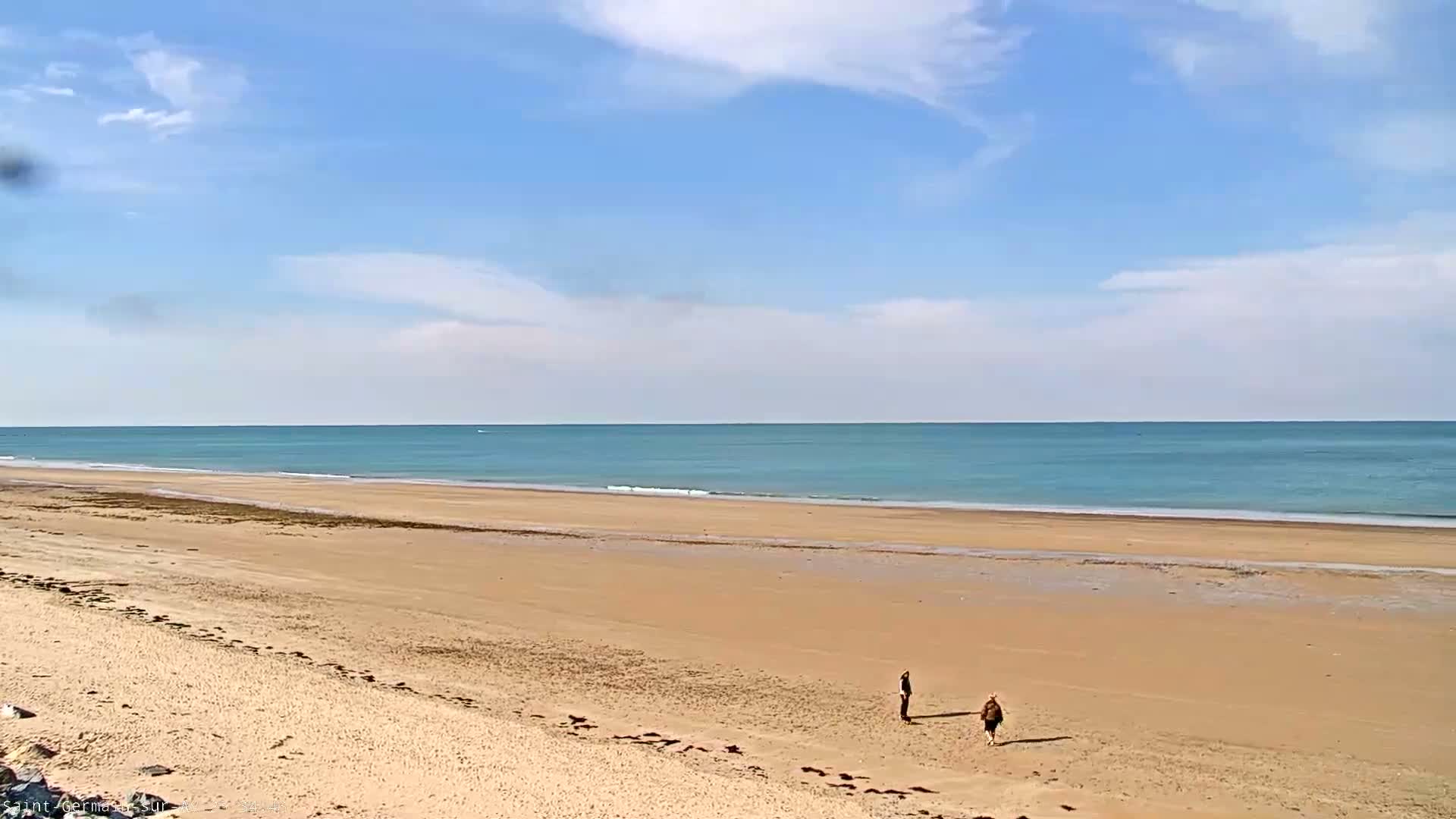 Under a mostly sunny sky, three people walk on a wide, sandy beach toward a calm, light-blue ocean.