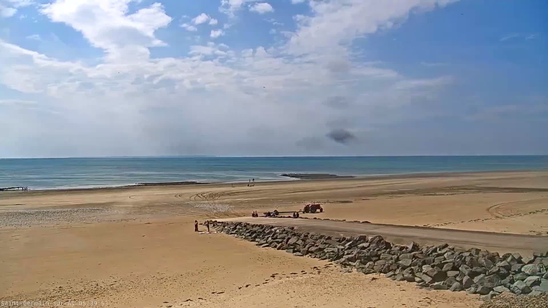 A sandy beach stretches to a calm ocean under a partly cloudy sky, with a tractor and several people visible near a rock wall.