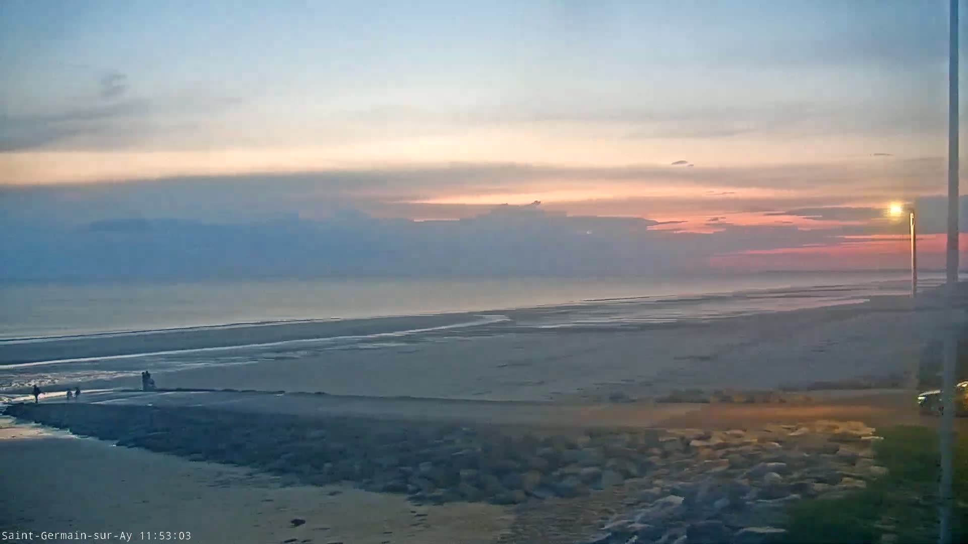 A pinkish sunset over a wide, sandy beach and calm ocean with a few people visible in the distance.