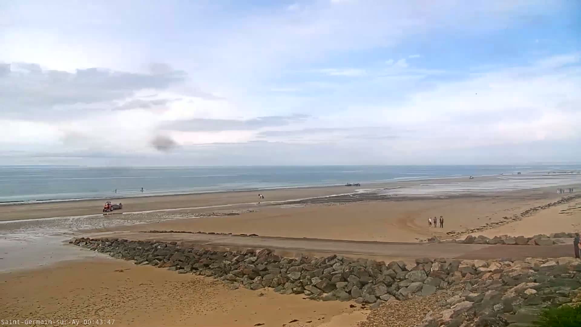 A tractor is on a wide, sandy beach next to a calm ocean under a partly cloudy sky.