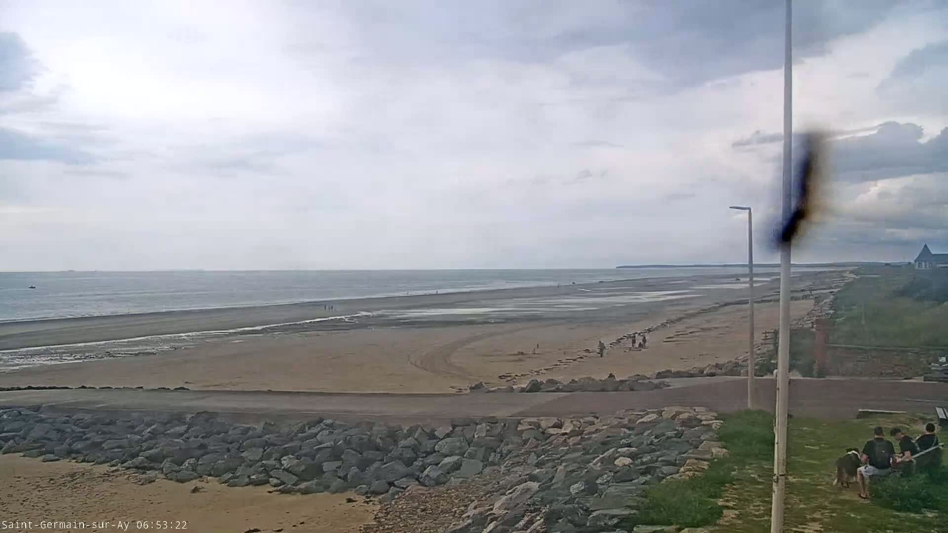 A wide sandy beach stretches to a calm ocean under an overcast sky, with a few people strolling along the shore and others sitting on a grassy area near a rock wall.