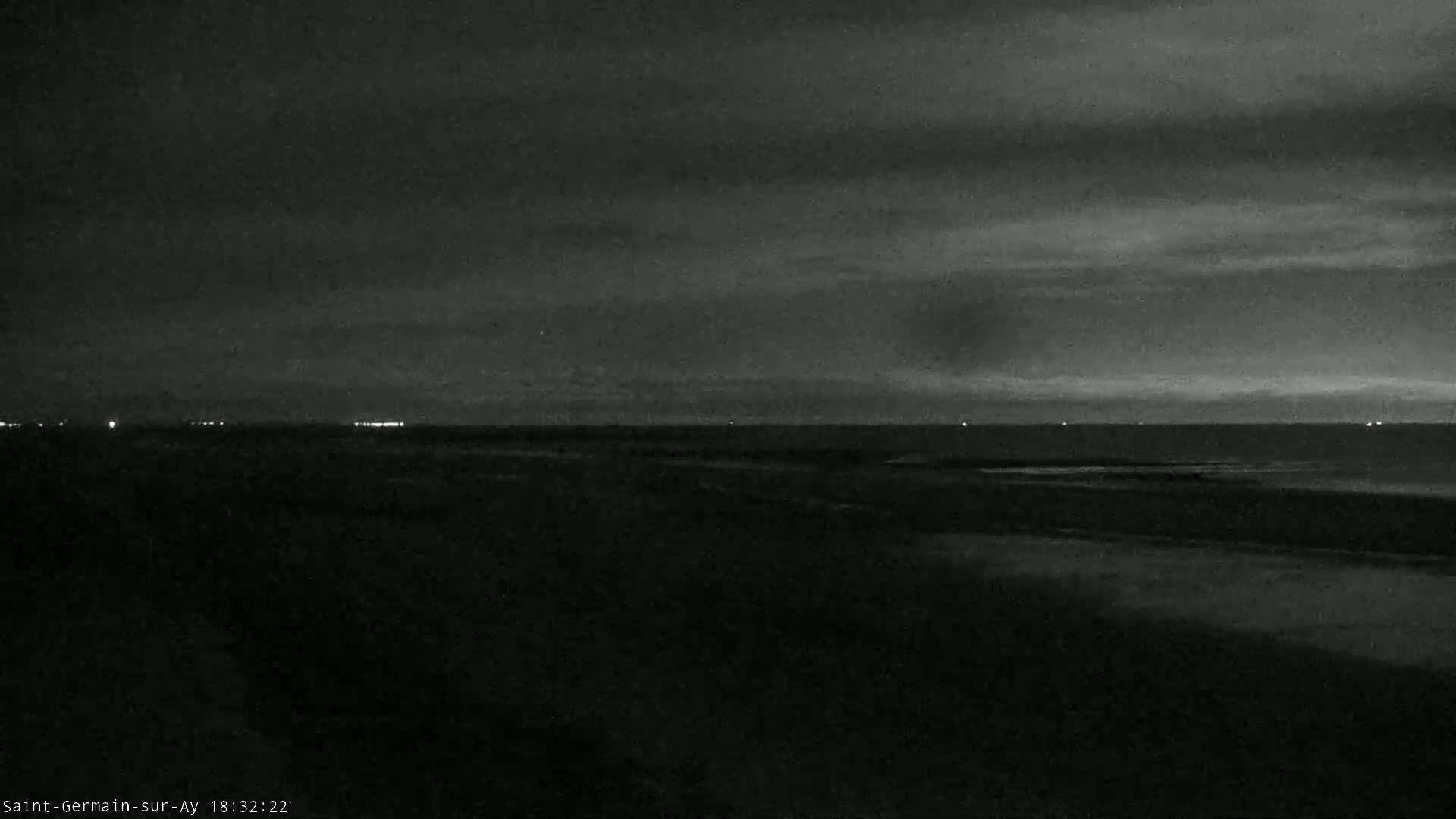 A dark, nighttime coastal scene shows a calm body of water under a cloudy sky with distant lights on the horizon.