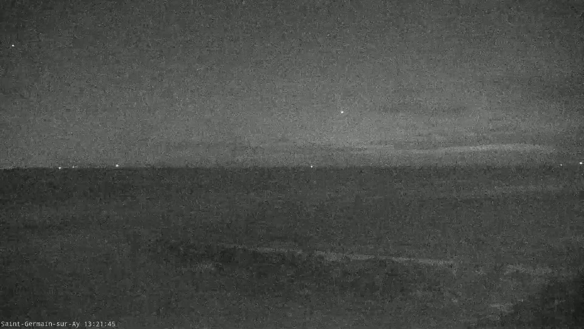 A low-light, nighttime image shows a flat, dark landscape under a mostly clear, dark sky with a few distant lights visible.